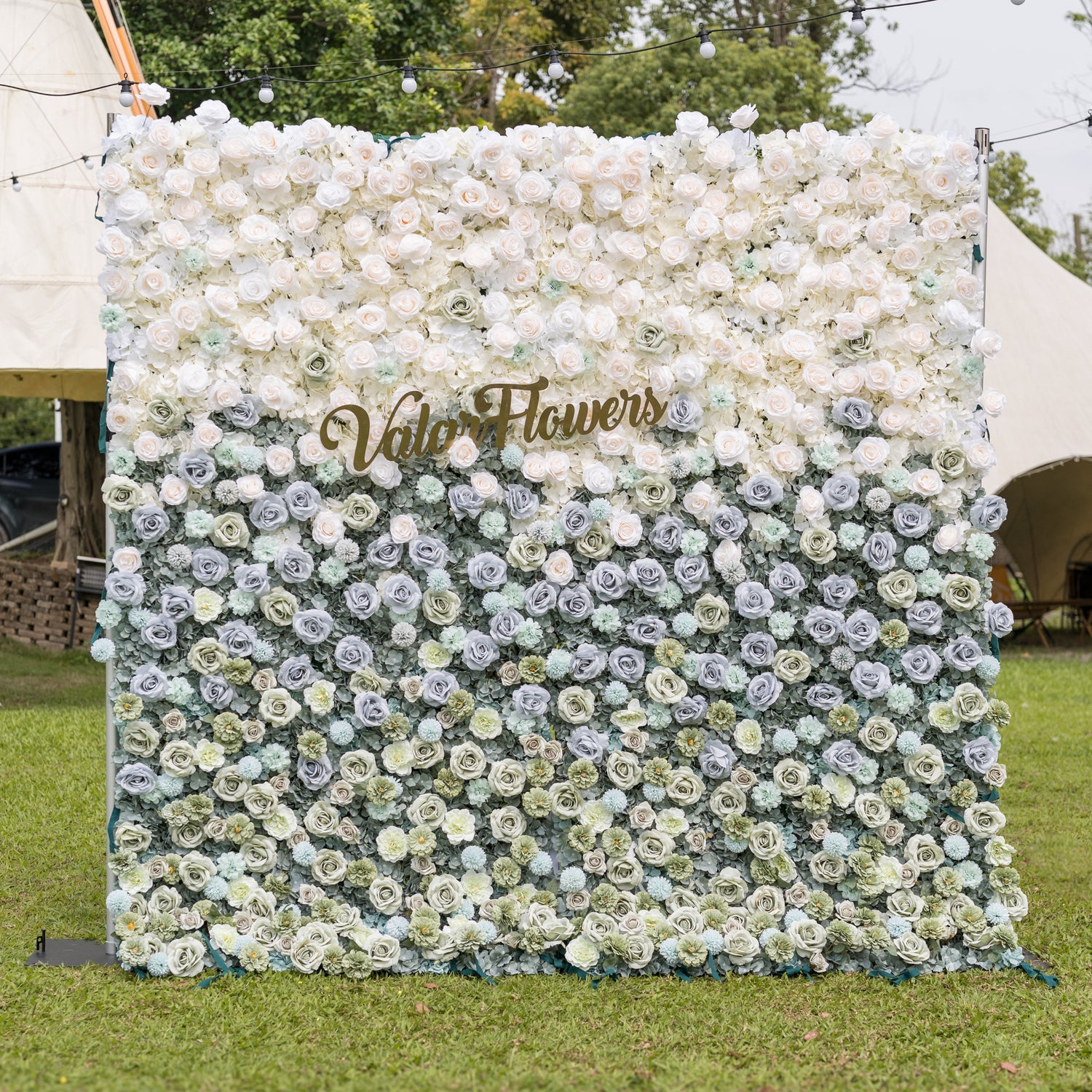 Valar Flowers Ethereal Serenity flower wall front view with dusty blue sage green and ivory roses dahlias and hydrangeas accented by a gold script sign on a green lawn under bistro lights for boho-chic weddings and elegant event backdrops