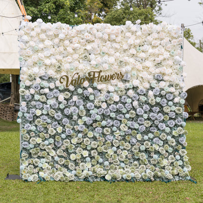 Valar Flowers Ethereal Serenity flower wall front view with dusty blue sage green and ivory roses dahlias and hydrangeas accented by a gold script sign on a green lawn under bistro lights for boho-chic weddings and elegant event backdrops