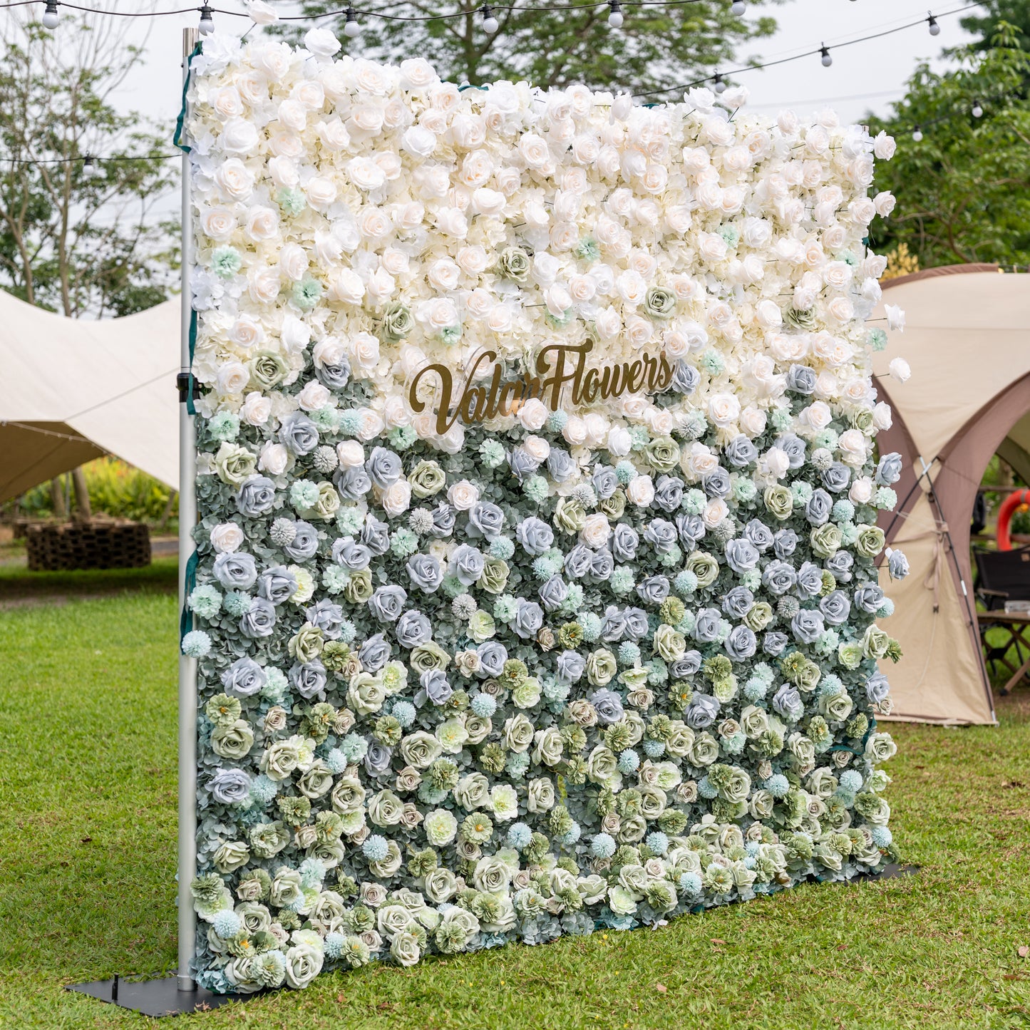 Three-quarter angle of Valar Flowers Ethereal Serenity artificial flower wall showcasing muted blue sage green and cream roses with silver eucalyptus foliage outdoors against canvas tents and globe lights for modern garden wedding ceremonies