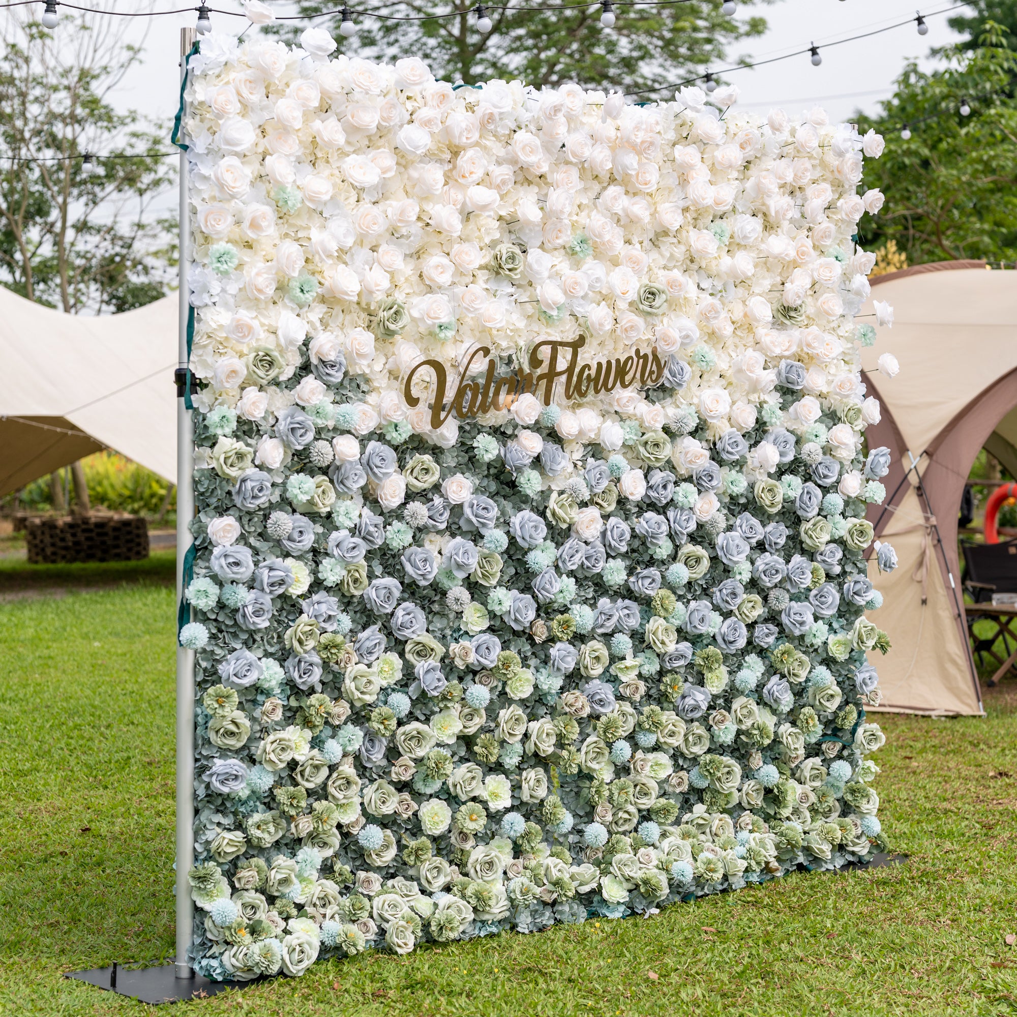 Three-quarter angle of Valar Flowers Ethereal Serenity artificial flower wall showcasing muted blue sage green and cream roses with silver eucalyptus foliage outdoors against canvas tents and globe lights for modern garden wedding ceremonies