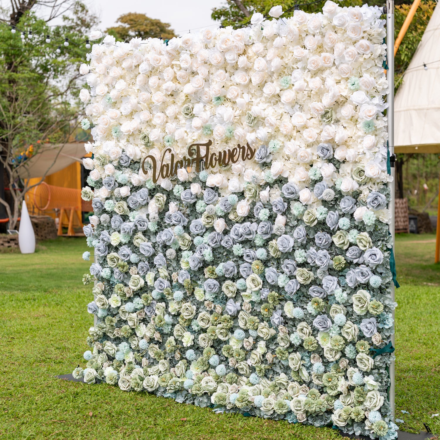 Valar Flowers Ethereal Serenity wall straight-on over manicured lawn beneath string lights featuring layered dusty blue ivory and sage blooms with a gold cursive Valor Flowers sign for romantic bridal shower and baby shower backdrops