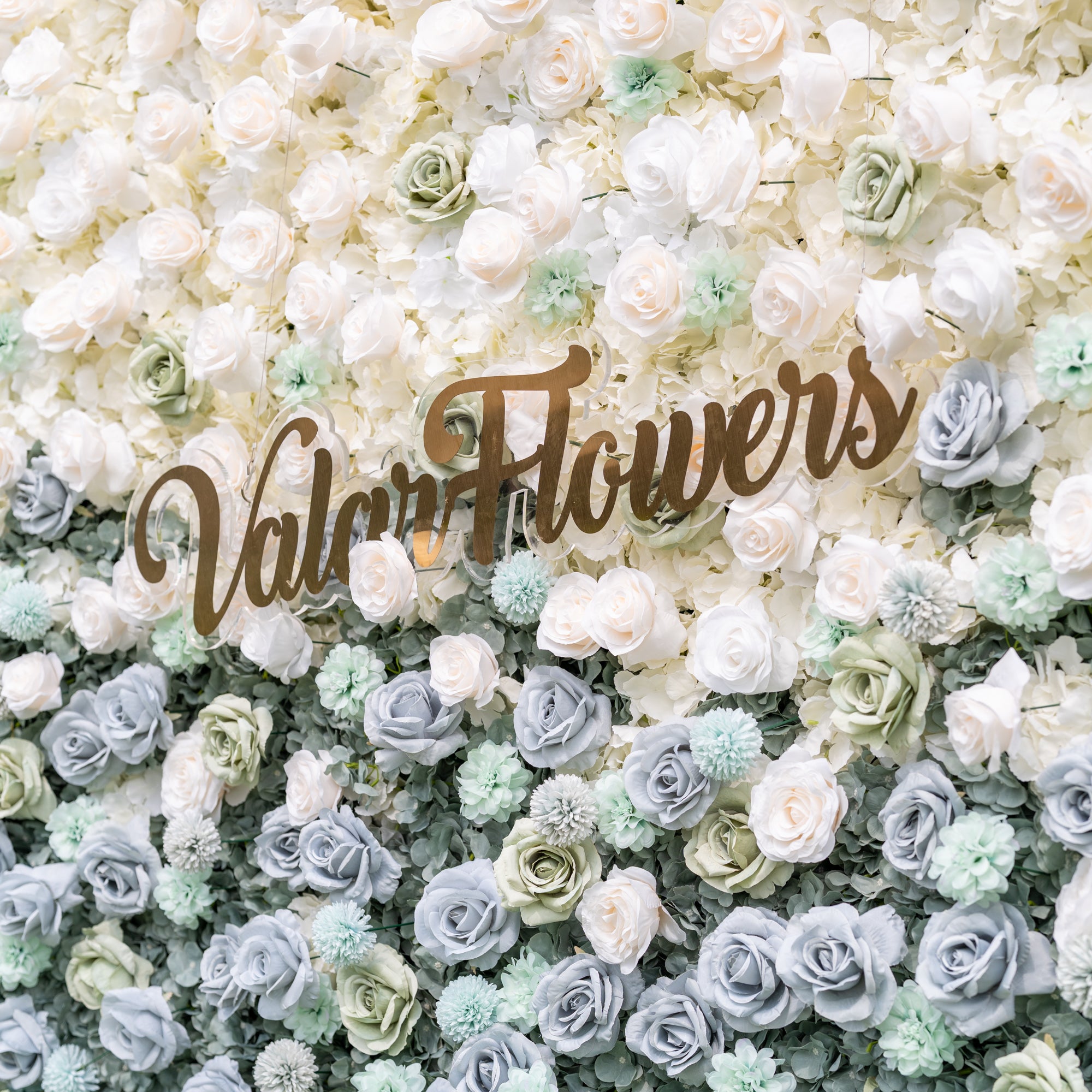 Close-up of Valar Flowers Ethereal Serenity flower wall highlighting dusty blue hybrid tea roses sage green pom-pom dahlias and creamy ivory hydrangeas with soft blurred outdoor backdrop for upscale event decor and photo booth installations