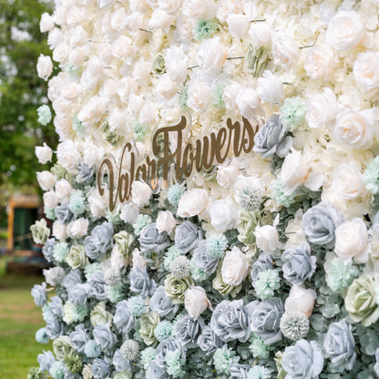 Low-angle close-up of Valar Flowers artificial flower wall emphasizing the sculptural depth of steel blue roses mint green pom-poms and ivory hydrangeas on fabric base with dreamy defocused garden background for bohemian wedding ceremonies