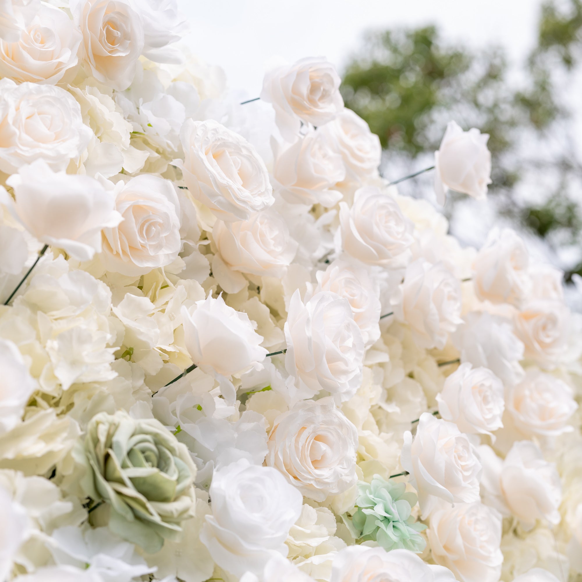 Angled close-up of Valar Flowers wall with gold cursive Valor Flowers sign among dusty blue and sage green roses against soft outdoor greenery an elegant branded focal point for modern weddings and upscale corporate event photo opportunities