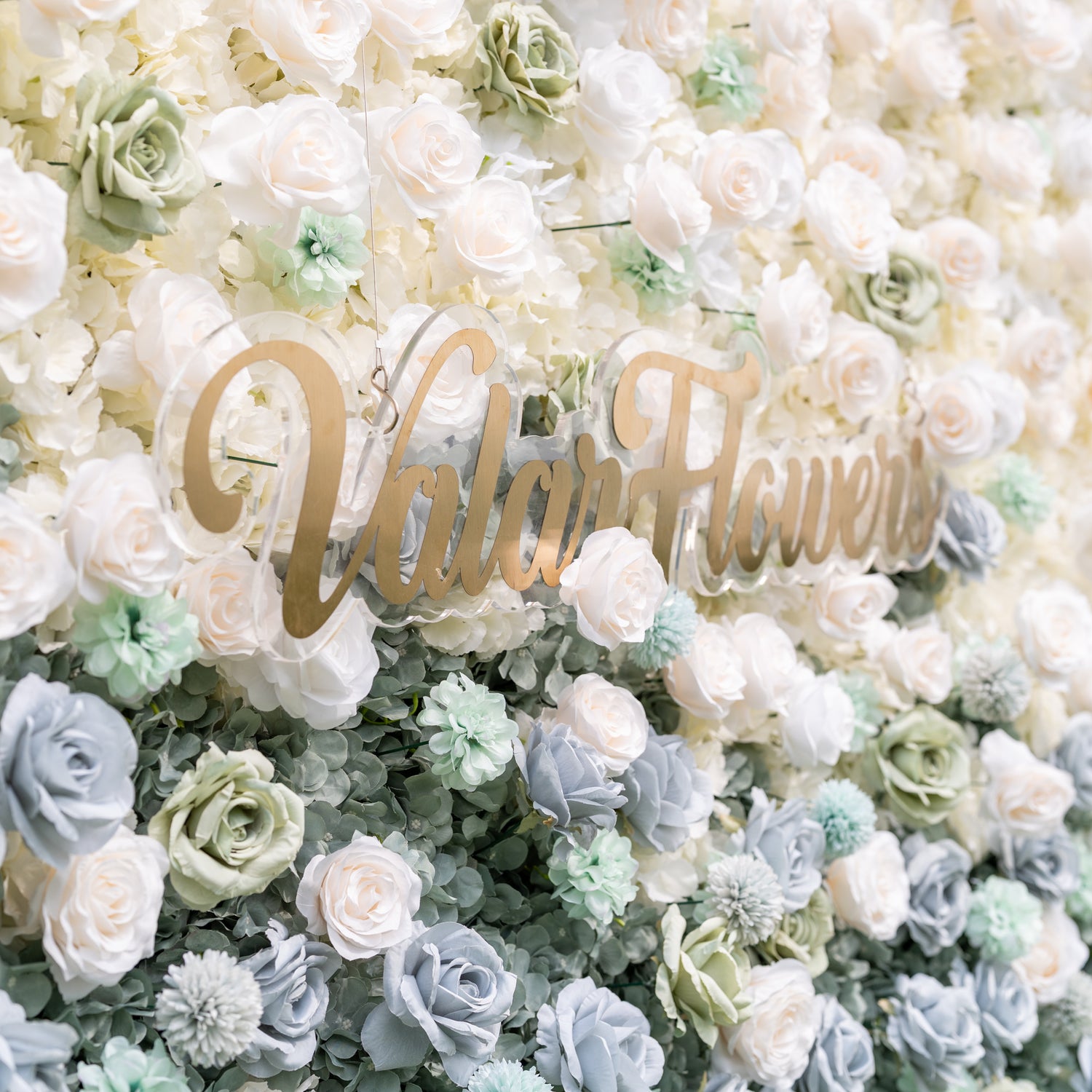 Dreamy close-up of Valar Flowers silk flower wall with dusty blue sage green and ivory roses accented by textured pom-pom blooms and eucalyptus sprigs in soft romantic blur evoking fine art for bridal portraits and luxury ceremony backdrops