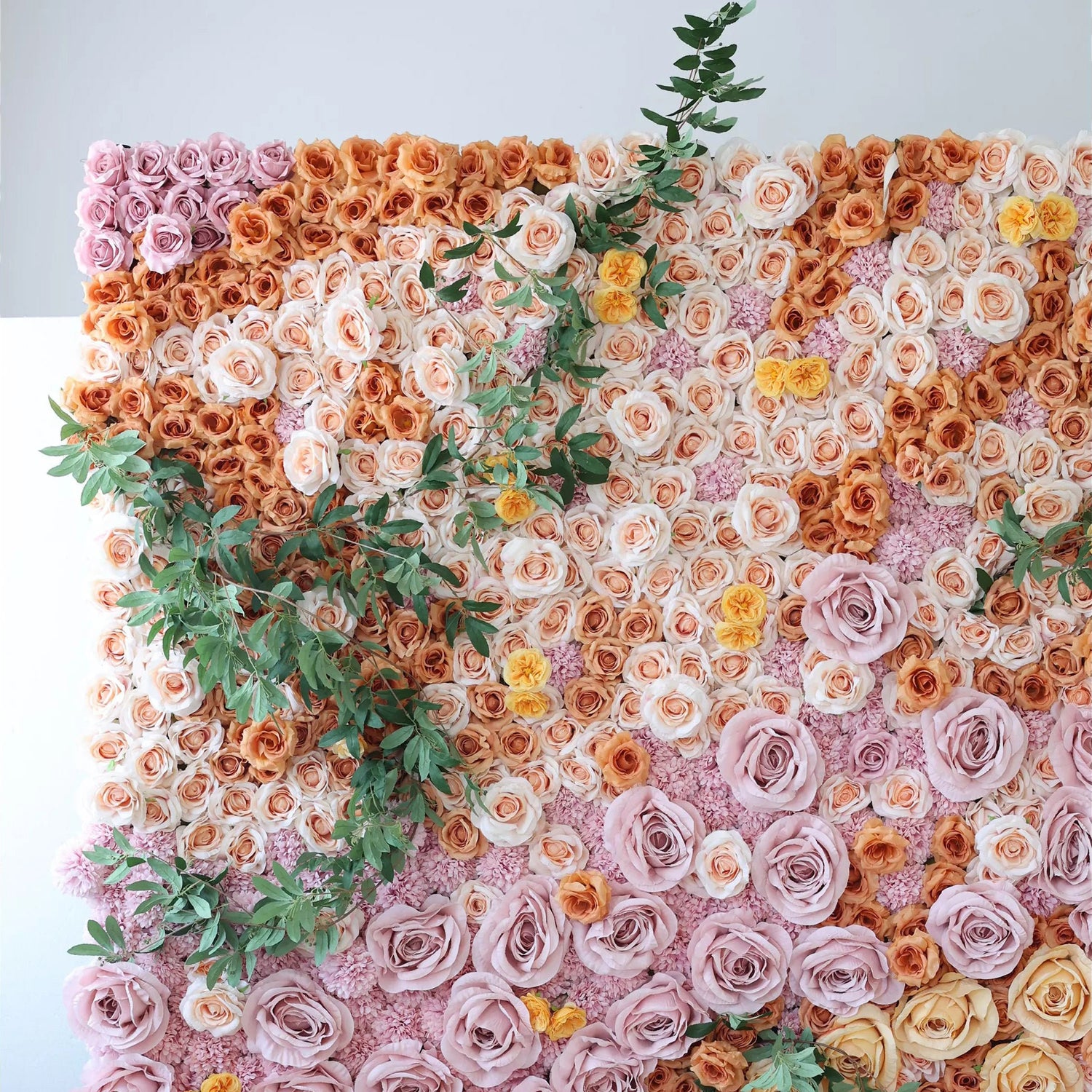 This image showcases a stunning floral wall by Valar Flowers. It features a dense, artful arrangement of artificial roses in a harmonious palette of soft pinks, warm oranges, and creamy whites, interspersed with delicate green foliage. The flowers create a lush, textured, and visually captivating display, exuding elegance and charm, perfect for adding a touch of sophistication to events or interior spaces.