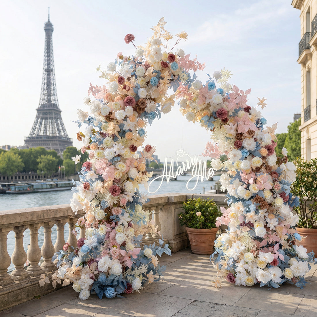 A dreamy, circular floral arch from Valar Flowers, bursting with a pastel palette of blush pink, sky blue, creamy white, and soft peach blooms—including roses, peonies, and delicate foliage—set against the iconic Eiffel Tower in Paris. A "Marry Me" sign rests at its center, creating a romantic, unforgettable atmosphere perfect for a proposal or wedding celebration.