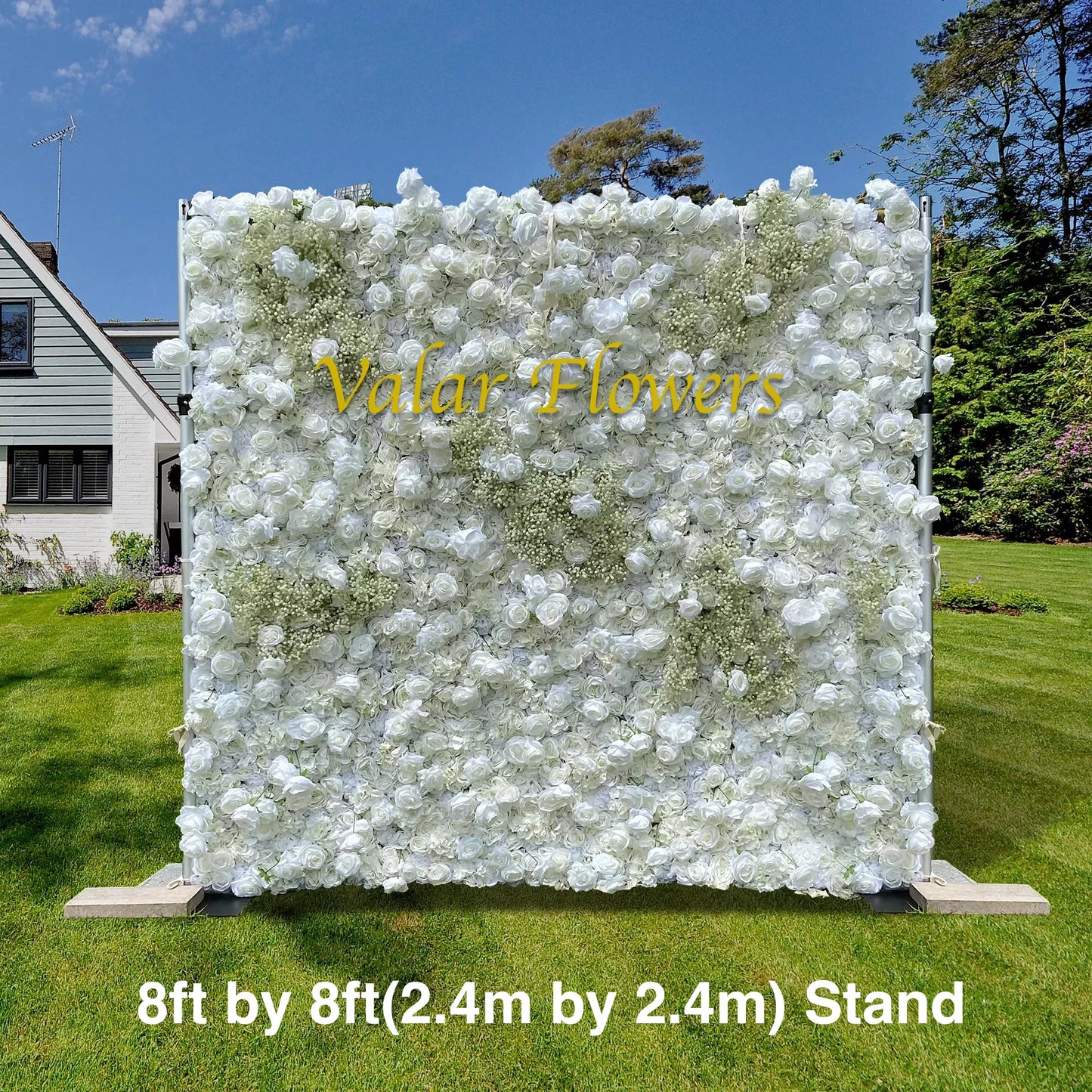 An 8ft by 8ft floral wall on a stand is covered in white artificial roses and small green accents. The text "Valar Flowers" is in yellow on the wall. It's set outdoors on a grassy area near a house. 
