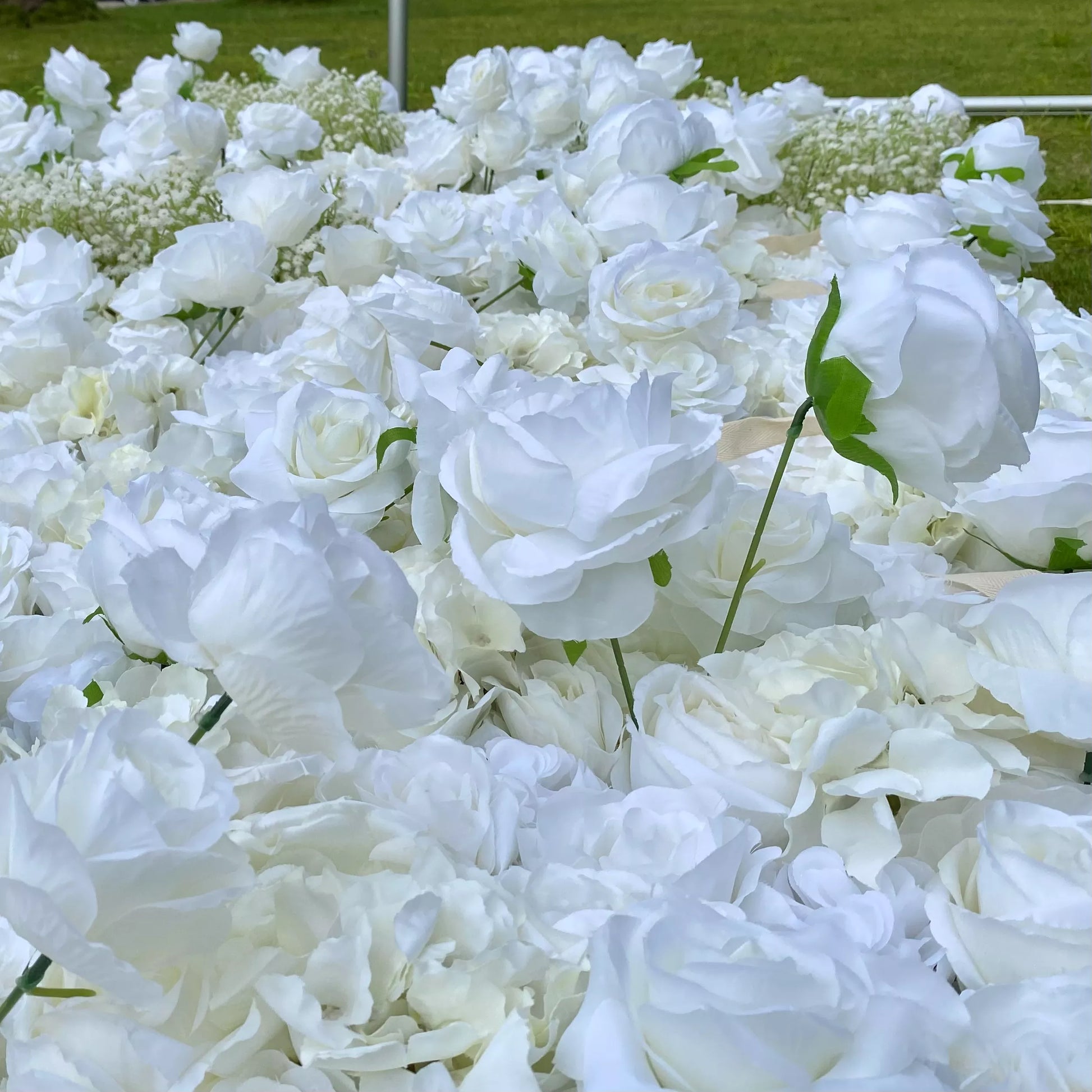 This image presents a dense cluster of white artificial flowers, likely roses, with lush petals. Some baby's breath is also visible in the background. Set outdoors on a grassy area, the pure white blooms create an elegant and serene atmosphere, perfect for decorative purposes in events or celebrations.