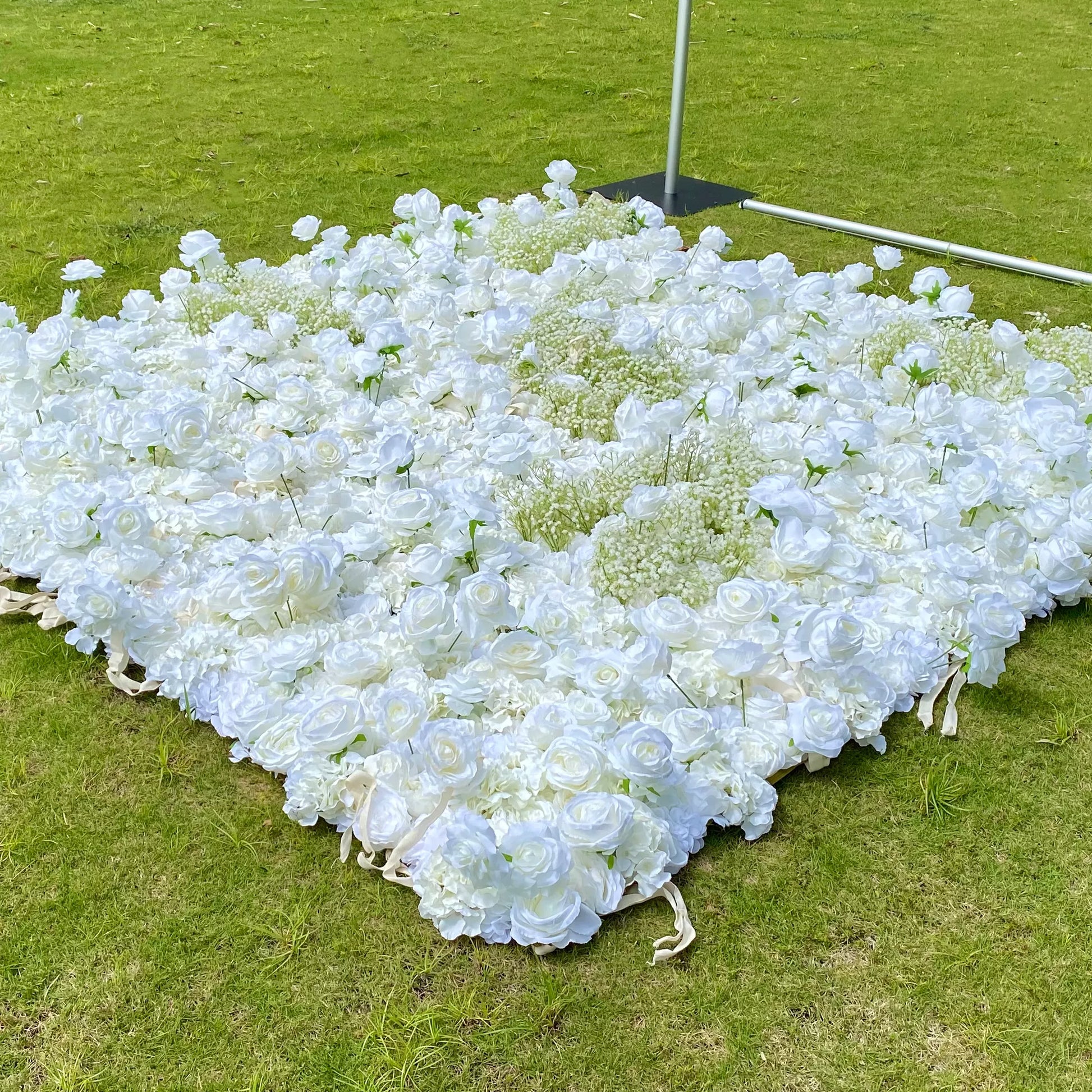 This image shows a large, elaborate arrangement of white flowers, likely roses and baby's breath, spread out on a lush green lawn. The flowers form a dense, elegant display, creating a soft and romantic look. Set outdoors under natural light, it gives off a serene and beautiful vibe, perfect for decorative or ceremonial purposes.