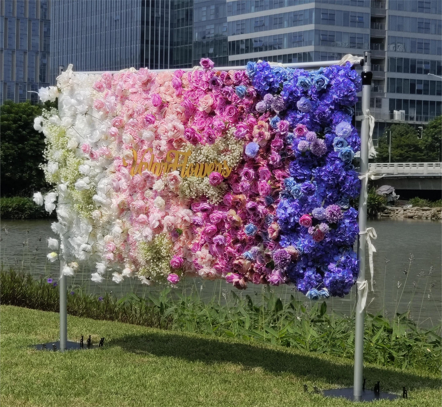 This photo showcases a vibrant floral wall with a gradient of white, pink, and blue flowers, including roses and hydrangeas, with baby's breath adding texture. "Valar Flowers" is in gold lettering. Set outdoors by a body of water with buildings in the background, it has a dreamy, romantic style, perfect for weddings or photo - ops. 
