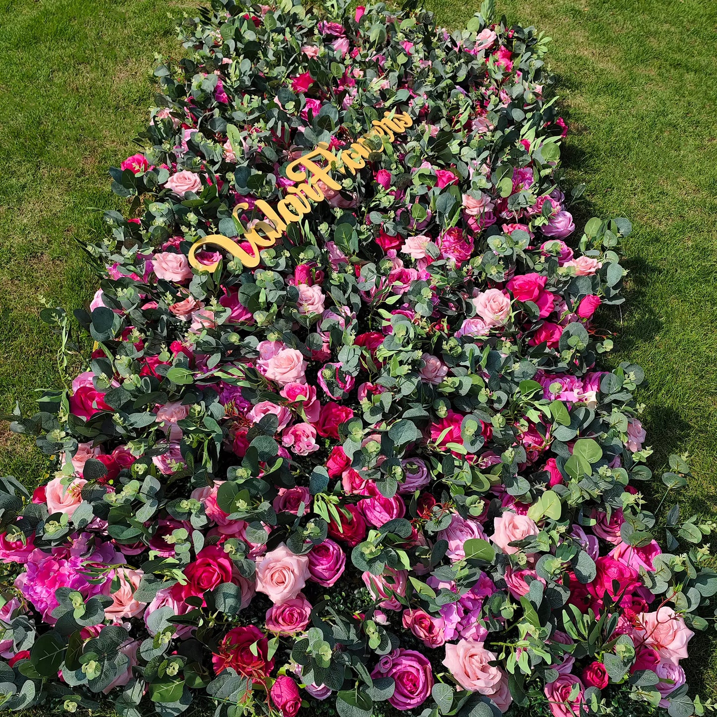 This image showcases a large, dense arrangement of artificial roses in shades of pink, red, and purple, interspersed with green eucalyptus leaves. At the center, there's a golden "Valar Flowers" sign. Set on a lush green lawn, the vibrant blend of colors and textures creates a visually appealing, elegant display, ideal for decorative purposes in events or as a striking floral installation.