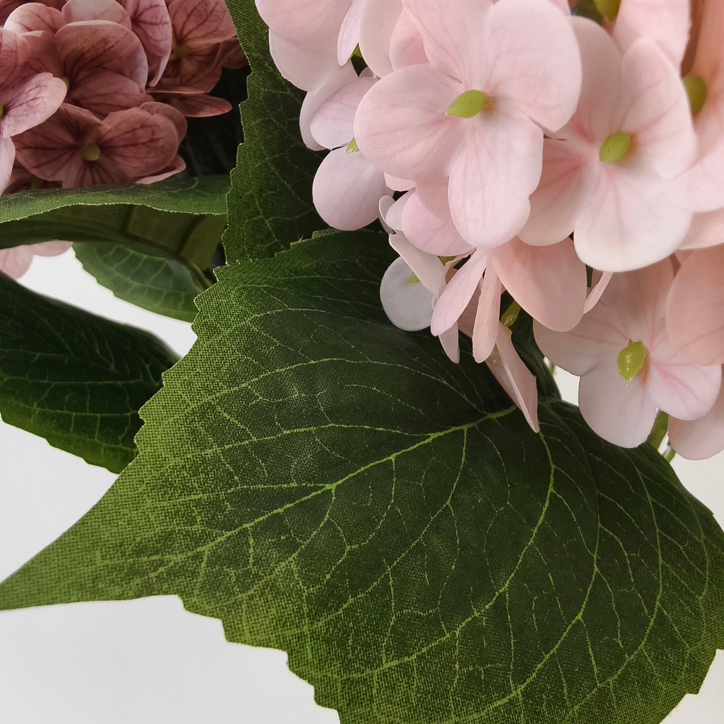 This close - up shows artificial hydrangea blooms. There are soft pink flowers with green centers and darker pink blossoms in the background. Large, textured green leaves with visible veins add realism. The arrangement has a delicate, romantic look, ideal for decorative displays. 