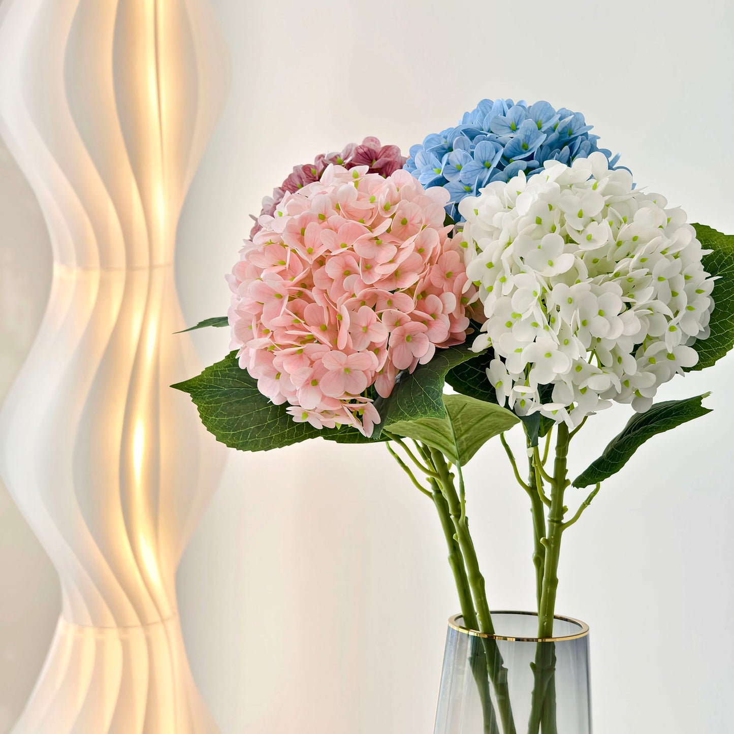 This arrangement features four hydrangea blooms in soft pink, deep pink, sky blue, and pure white, with lush green leaves. Placed in a sleek glass vase with a gold rim, it brings a fresh, elegant touch to home decor, blending pastel hues for a charming, serene aesthetic. 