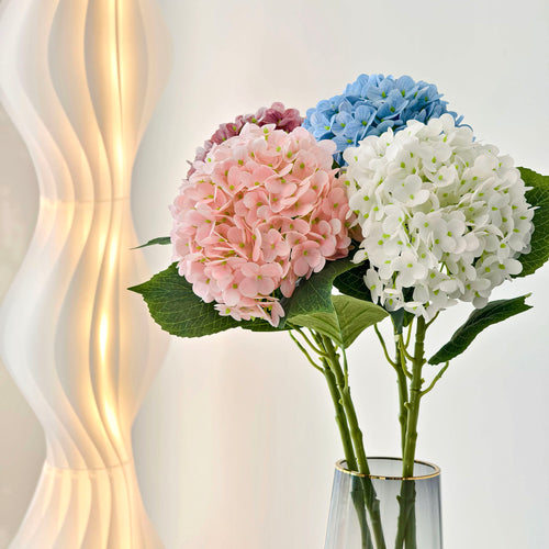 This arrangement features four hydrangea blooms in soft pink, deep pink, sky blue, and pure white, with lush green leaves. Placed in a sleek glass vase with a gold rim, it brings a fresh, elegant touch to home decor, blending pastel hues for a charming, serene aesthetic. 