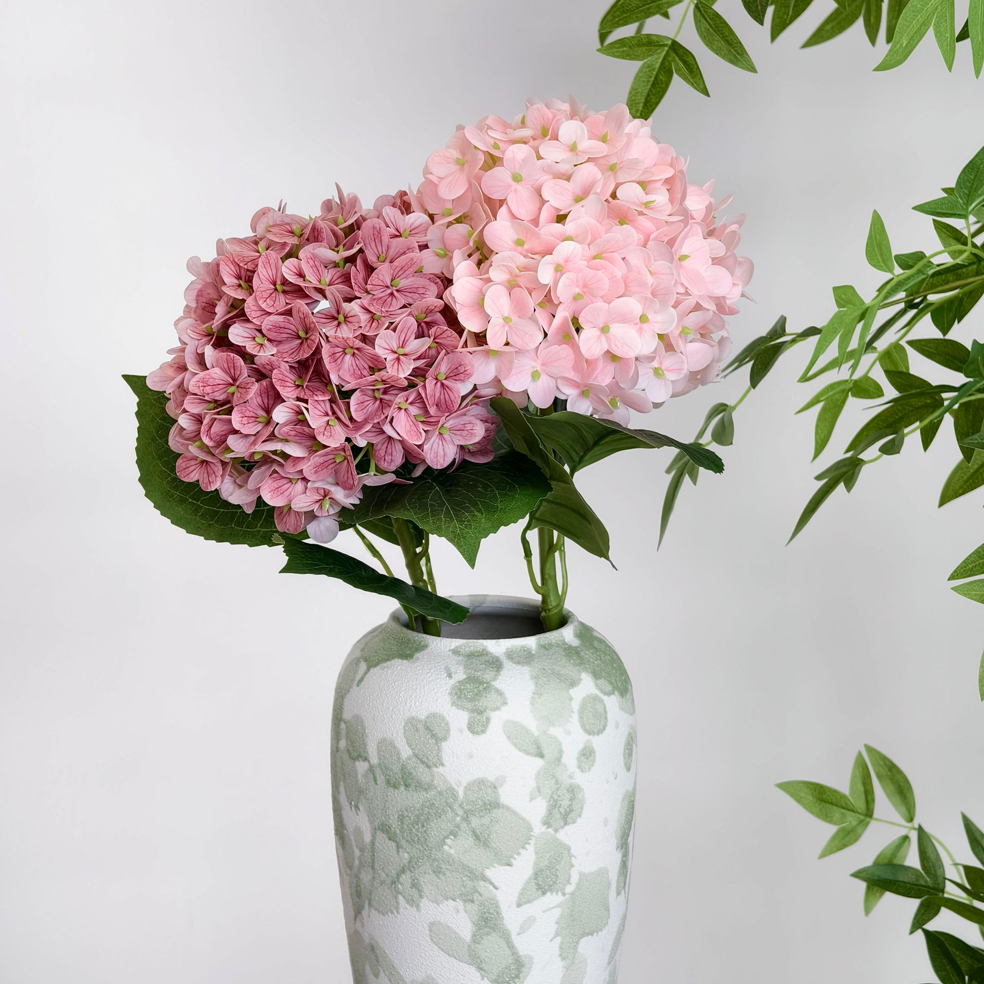 This arrangement features two artificial hydrangea blooms—one in deep pink, the other in soft pink—with green leaves, placed in a white vase with green watercolor - like patterns. The pastel hues and minimalist design create a gentle, elegant decor vibe, perfect for adding a touch of natural beauty to any space. 
