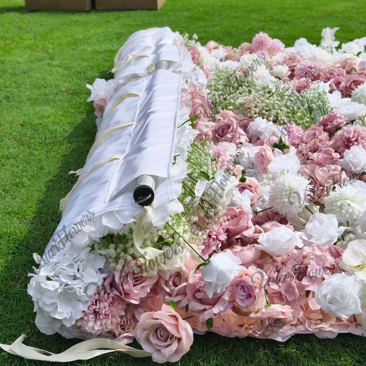 This image shows a floral arrangement laid on a grassy surface, likely part of a backdrop setup. The flowers include a mix of white and pink roses, white hydrangeas, and clusters of baby's breath. The color palette features soft pastels, with various shades of pink and white. The style is romantic and delicate, evoking a gentle and elegant atmosphere, suitable for events like weddings or garden - themed parties. The white fabric with gold - toned accents appears to be part of the structural support for the 