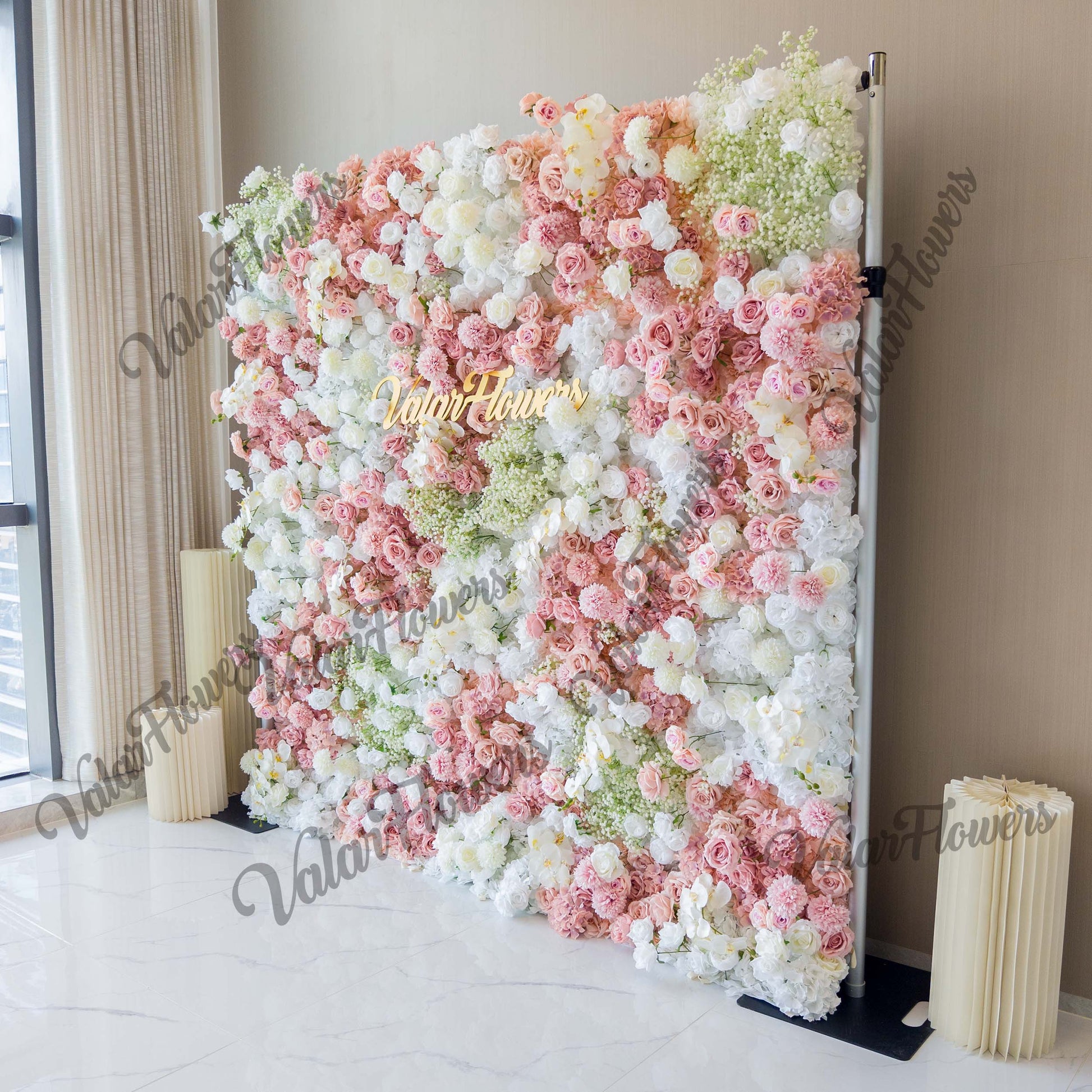This backdrop is a rectangular floral wall supported by metal poles. It features a mix of artificial roses in soft pink, light peach, and white hues, along with clusters of baby's - breath - like flowers. The color palette is gentle and romantic, with various pastel shades. The style is lush and elegant, perfect for wedding or event decor. The "ValarFlowers" sign in gold adds a touch of branding. 