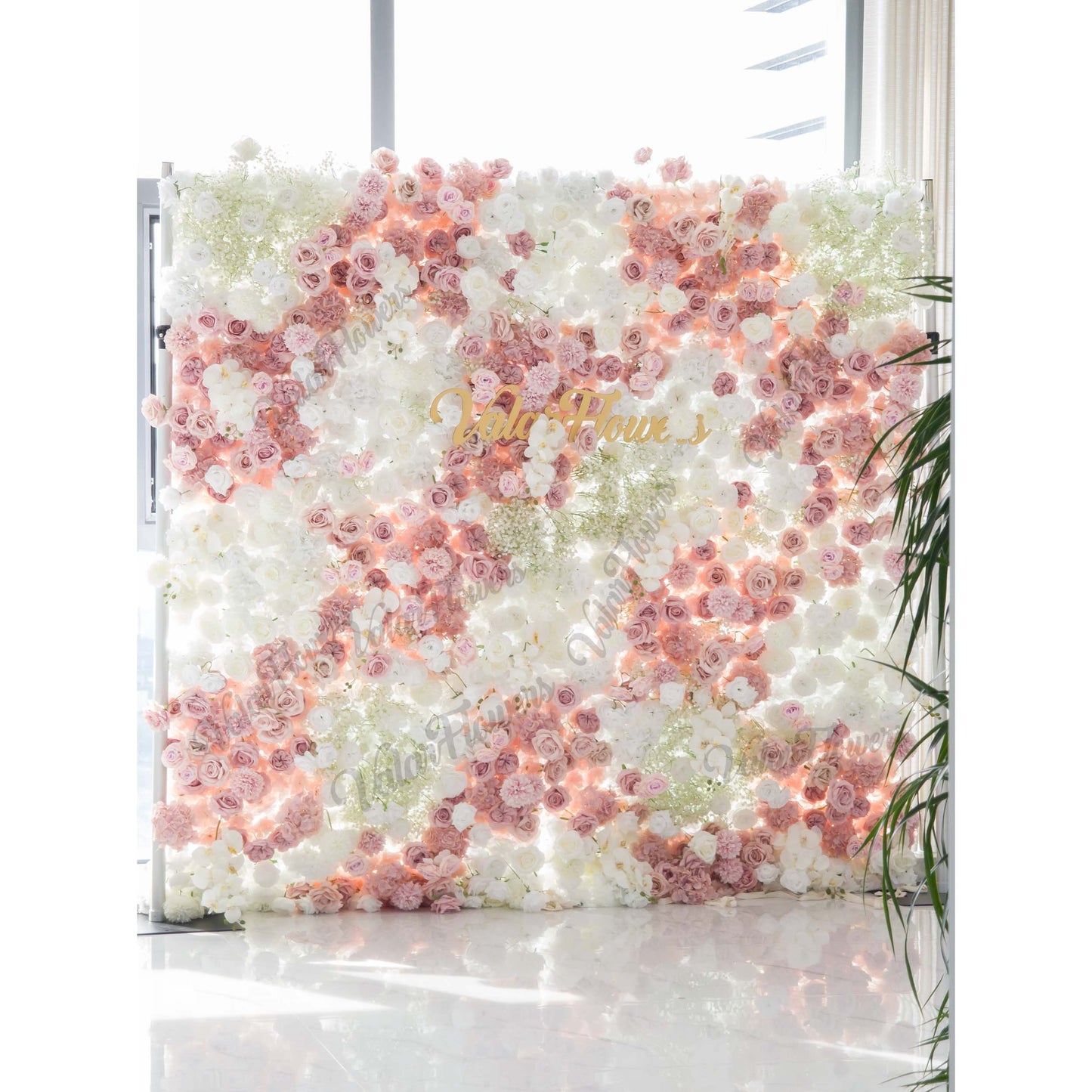 This backdrop is a rectangular floral wall supported by metal frames. It is densely covered with artificial flowers, including roses in shades of pink and white, along with clusters of baby's breath. The color palette consists of soft pinks, whites, and hints of green. The style is romantic and elegant, perfect for wedding - related events. The "ValarFlowers"  sign in gold adds a branded element. 