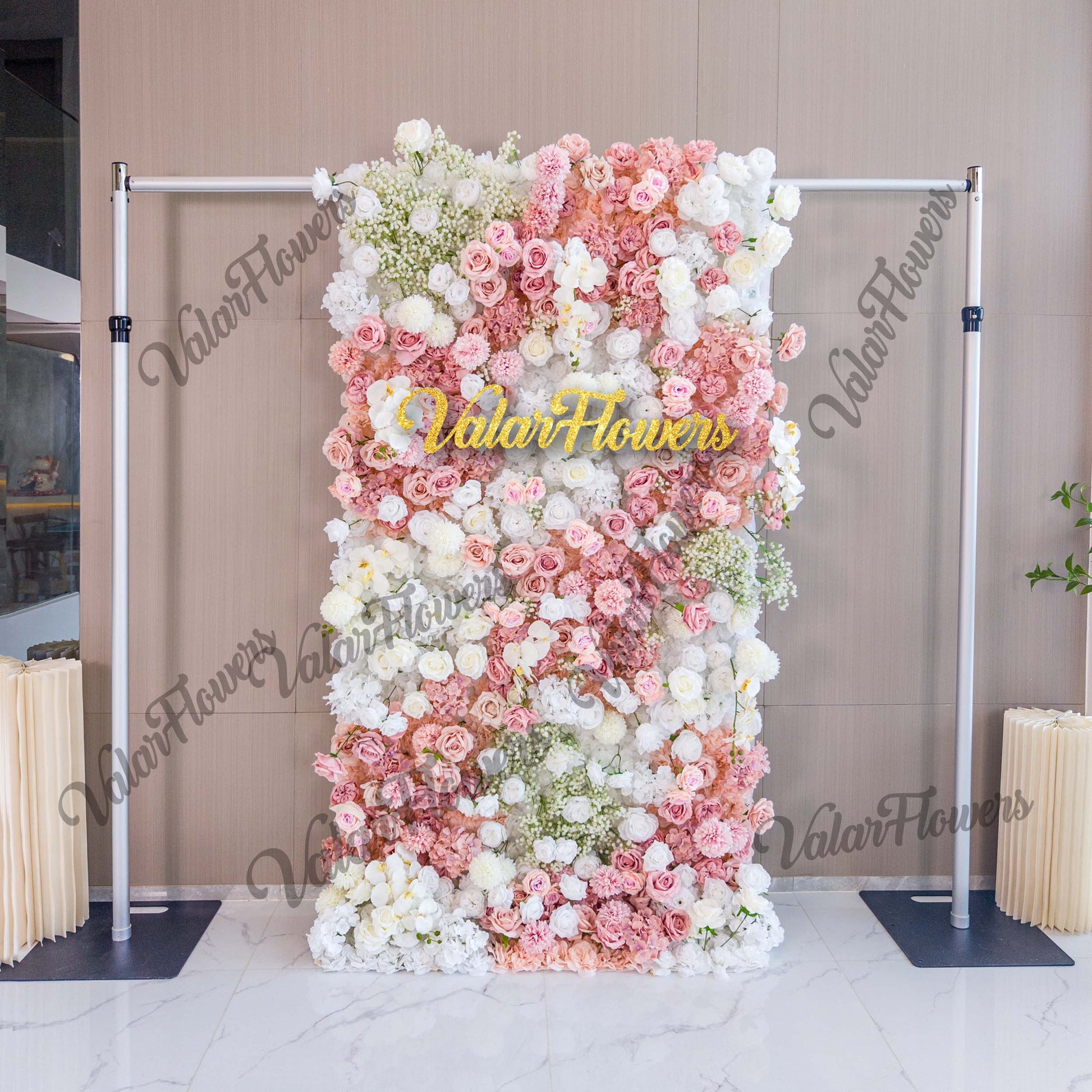 This backdrop is a vertical floral wall supported by a metal frame. It is densely decorated with artificial flowers, including roses in various shades of pink and white, along with clusters of baby's breath. The color palette features soft pastels, creating a romantic and elegant style, perfect for wedding or event settings. The "ValarFlowers" sign in gold adds a branded touch. 