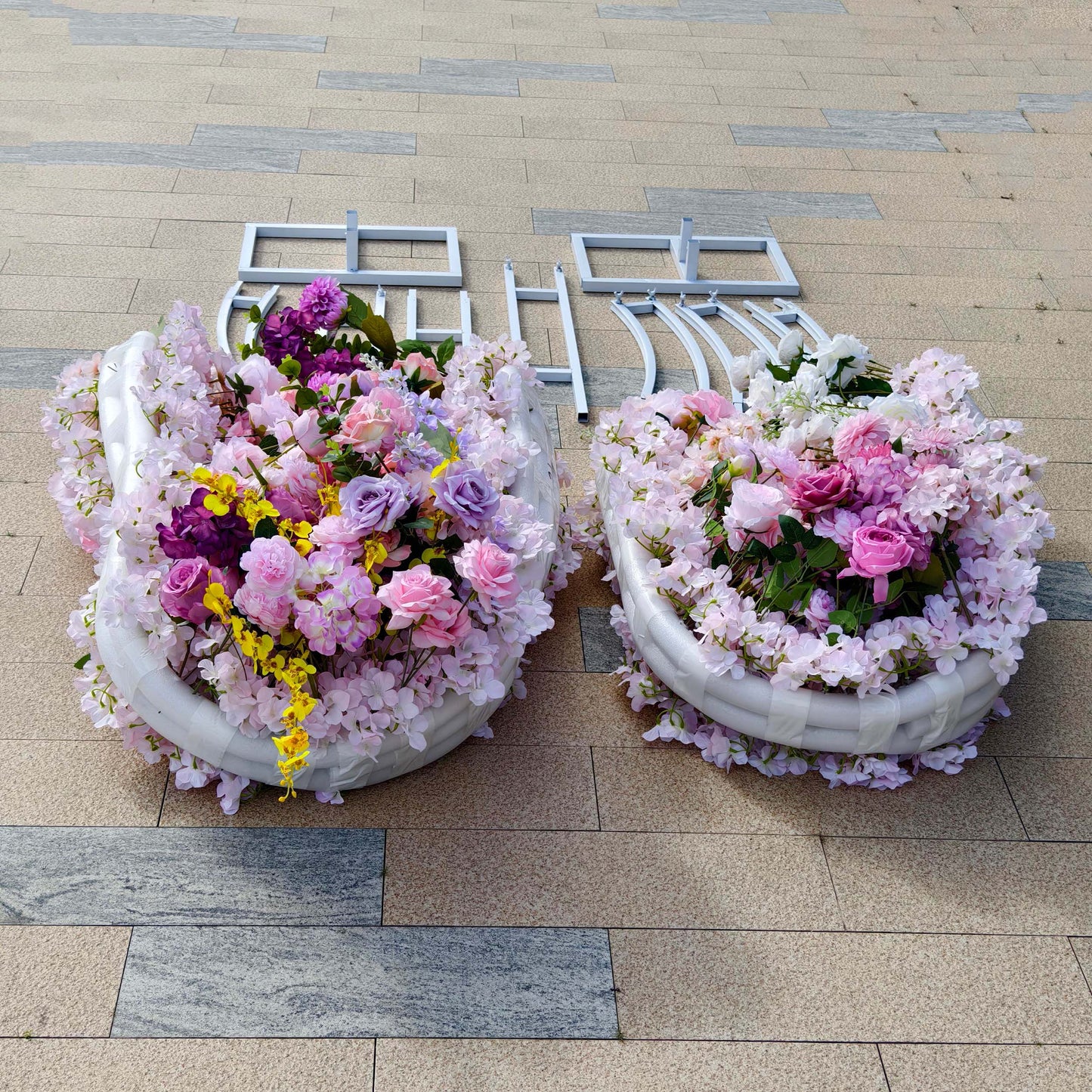 This Valar Flowers floral arch kit features two lush, curved arrangements of blush pink, lavender, and white blooms—including roses, hydrangeas, and wisteria—interwoven with vibrant yellow accents. The set includes a white metal frame for assembly, creating a romantic, versatile setup ideal for weddings, engagement shoots, or elegant outdoor celebrations.