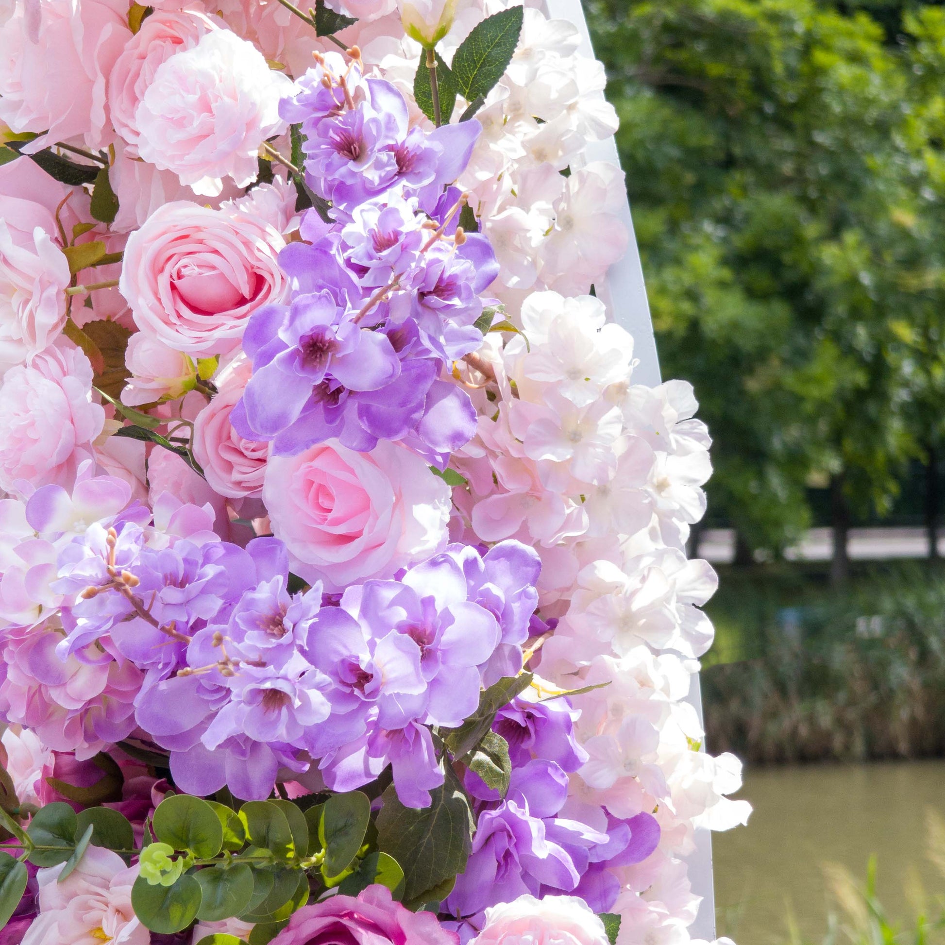 This close-up from Valar Flowers features a lush, romantic cluster of soft pink roses, vibrant purple orchids, and delicate white blooms, interwoven with glossy green foliage. Set against a serene outdoor backdrop of water and trees, the rich textures and fresh colors create a dreamy, sophisticated atmosphere, ideal for outdoor weddings, engagement shoots, or elegant celebrations.