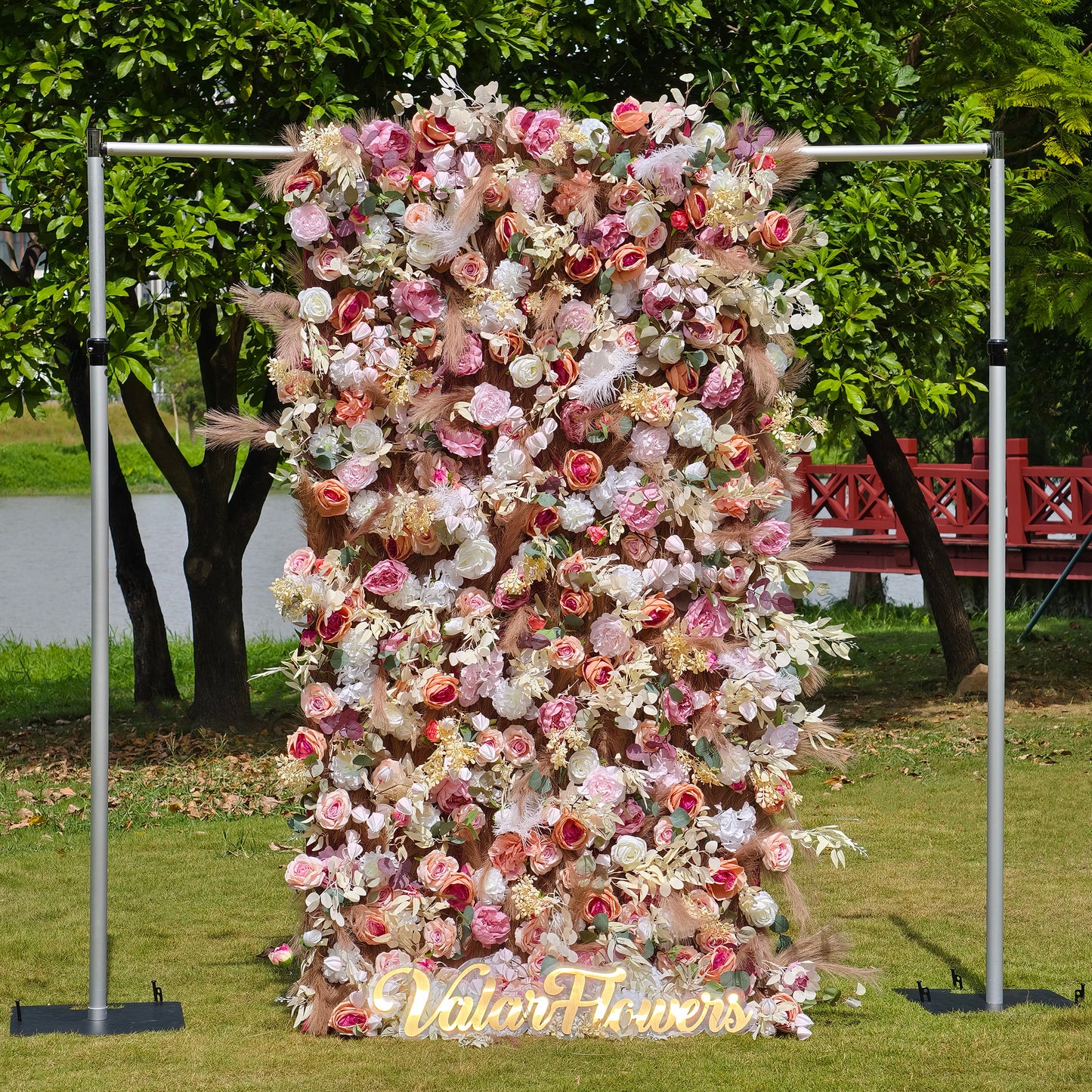 This photo showcases a large, elaborate floral backdrop supported by metallic poles. It is densely adorned with an array of artificial roses, peonies, and other blooms in soft pink, peach, and white hues, interspersed with eucalyptus leaves and pampas grass. The sign “ValarFlowers” is prominently displayed. Ideal for weddings or garden parties, it creates a romantic and dreamy atmosphere. 