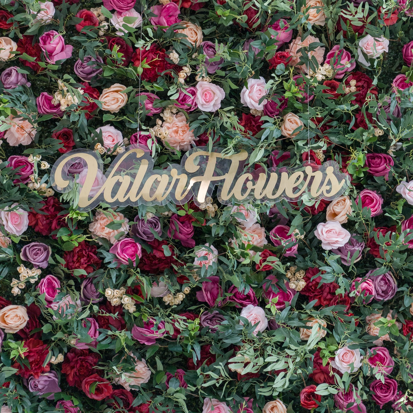 A lush artificial flower wall with pink, red, purple, and peach blooms, plus green foliage. At the center, a gold “ValarFlowers” sign on clear acrylic stands out. The vibrant, varied flowers create a romantic, decorative backdrop, perfect for events or branding displays. 