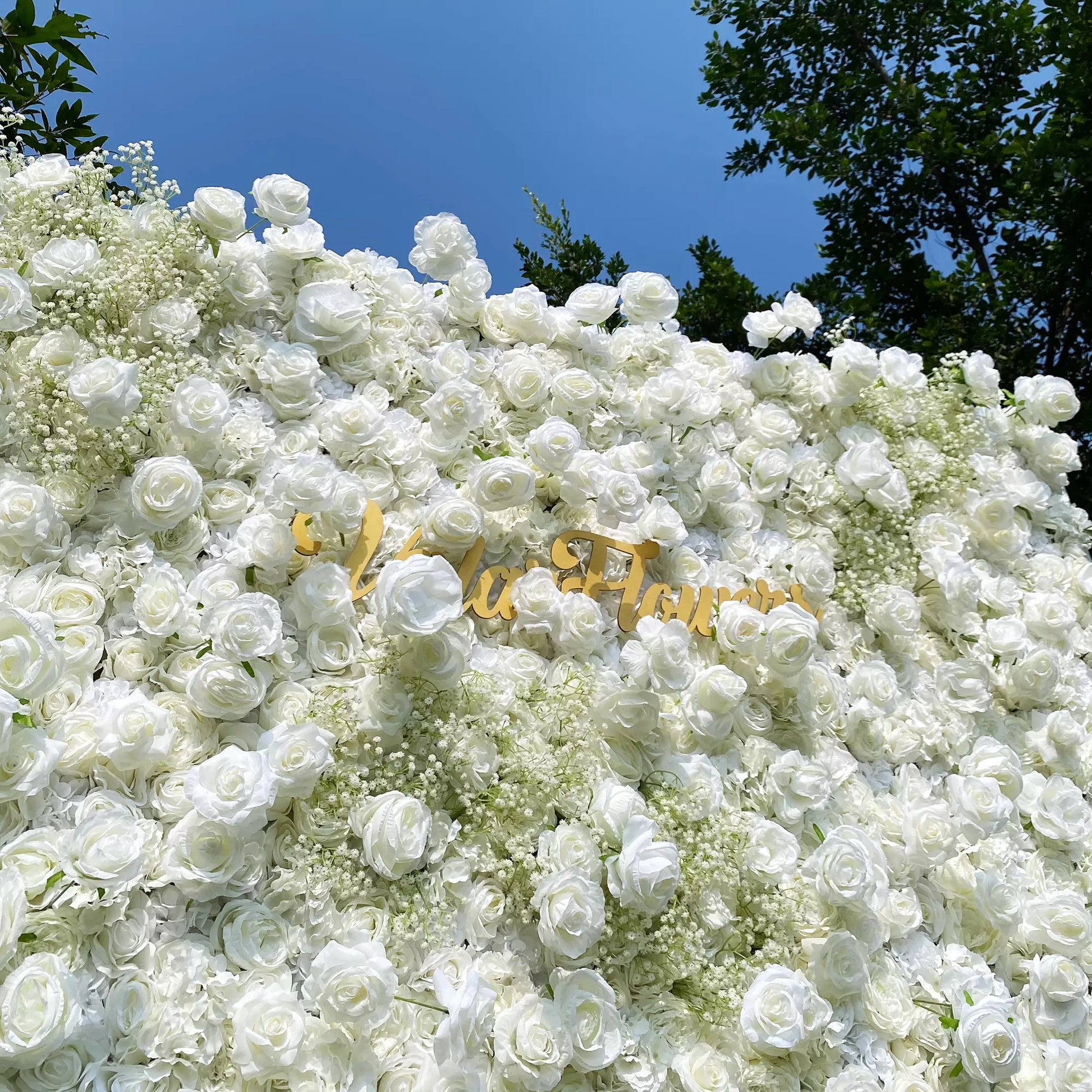 This image showcases a lush arrangement of white roses and baby's breath, forming a dense, elegant floral display. Golden text reading "Valar Flowers" is prominently featured. Set against a clear blue sky with tree branches in the background, it exudes a refined and romantic atmosphere, perfect for decorative or celebratory purposes.
