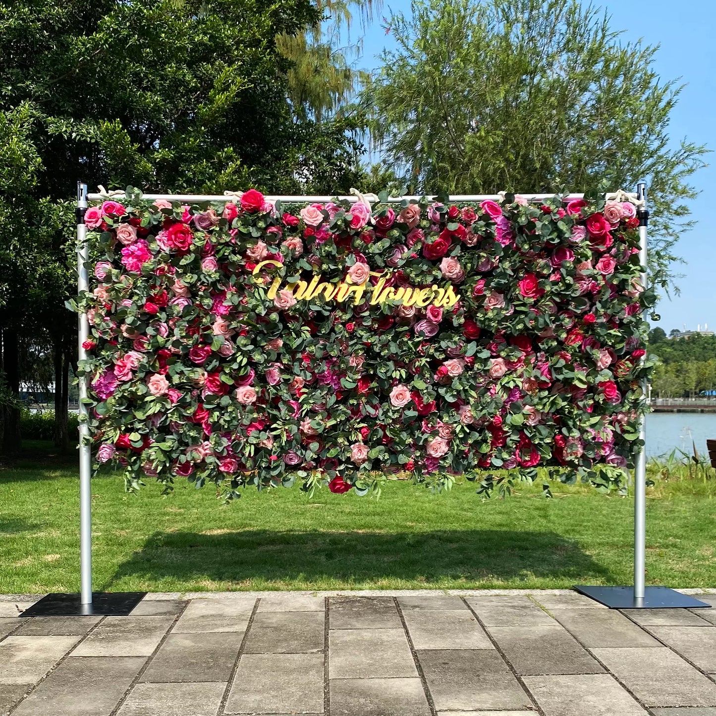 This image features a 4x8ft floral wall set outdoors. It's densely covered with roses in shades of pink, red, and purple, interspersed with green foliage. A golden "Valar Flowers" sign adorns the center. The backdrop includes a lawn, trees, a lake, and a clear blue sky, creating a vibrant, elegant scene perfect for events, exuding natural beauty and charm.