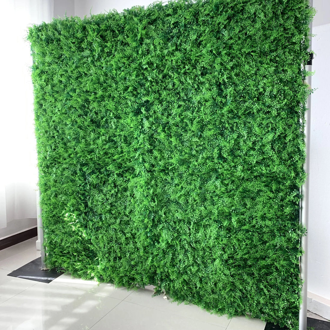 This image showcases a lush artificial greenery wall set indoors on a glossy white floor. It features dense, uniform green foliage with varied textures, creating a rich, natural, and visually refreshing display. The vibrant green hues and realistic plant details make it an ideal backdrop for adding organic elegance to interiors or events, exuding a serene and sophisticated atmosphere.