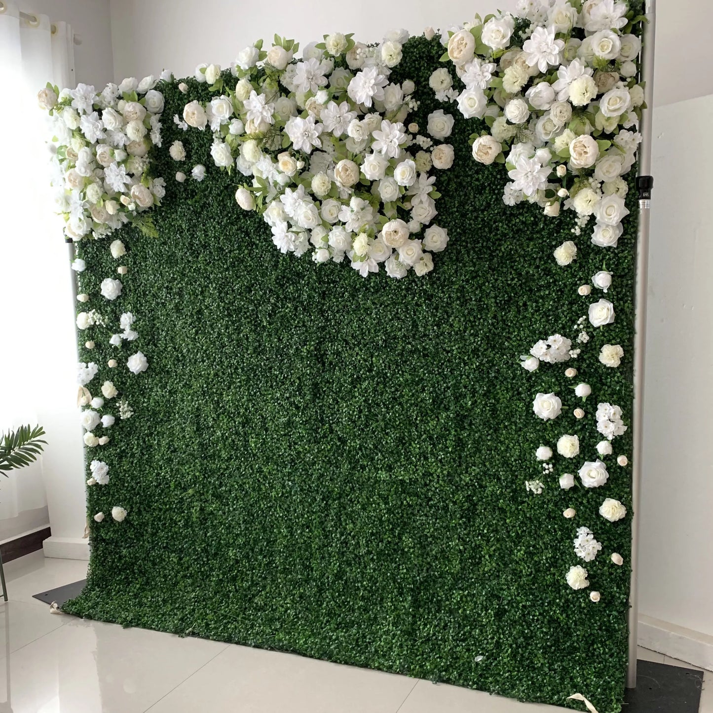 This elegant indoor floral wall from **Valar Flowers** features a lush, deep green foliage base, adorned with a cascading arrangement of white and cream blooms—including roses, hydrangeas, and delicate filler flowers. The blooms form a soft, heart-shaped design at the top, with individual flowers trailing down the sides. Set against a minimalist white backdrop, the design evokes a fresh, romantic, and naturally sophisticated atmosphere ideal for weddings, photo backdrops, or elegant celebrations.