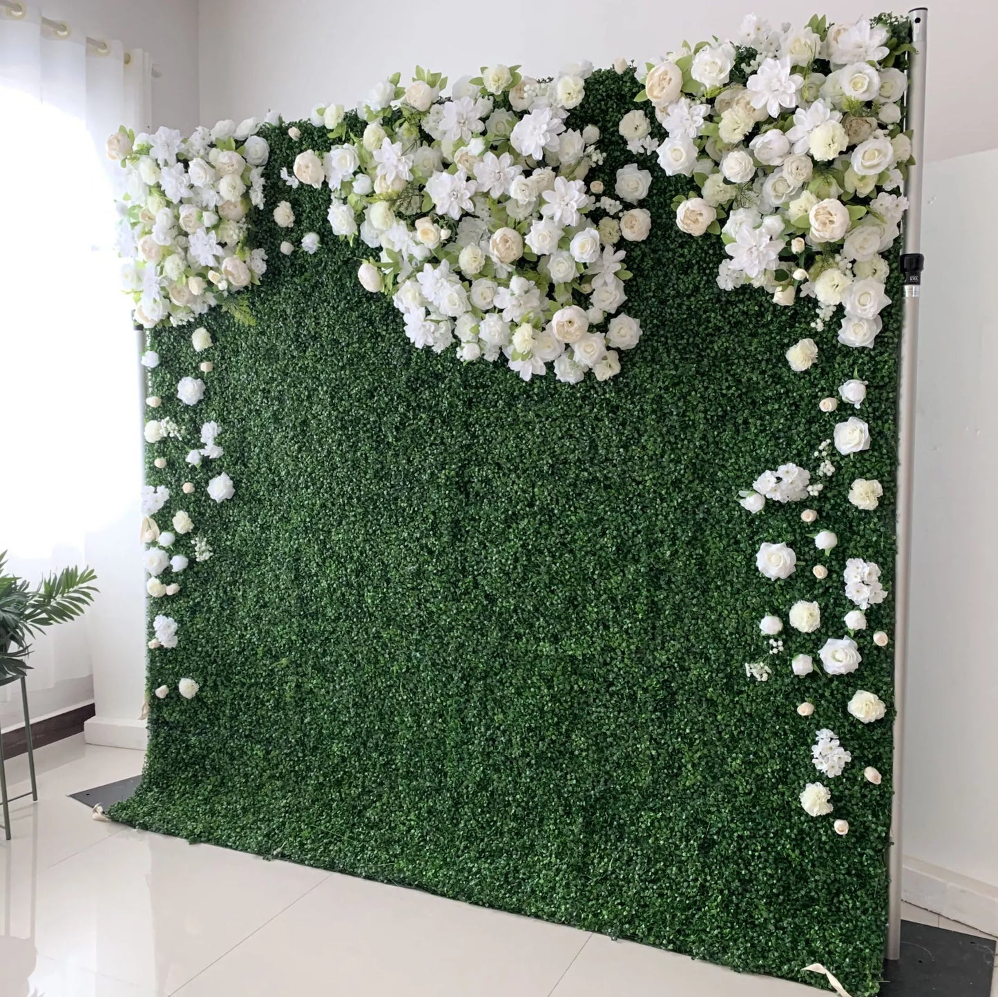 This elegant indoor floral wall features a lush, deep green foliage base, adorned with a cascading arrangement of white and cream blooms—including roses, hydrangeas, and delicate filler flowers. The blooms form a soft, heart-shaped design at the top, with individual flowers trailing down the sides. Set against a minimalist white backdrop, the design evokes a fresh, romantic, and naturally sophisticated atmosphere ideal for weddings, photo backdrops, or elegant celebrations.