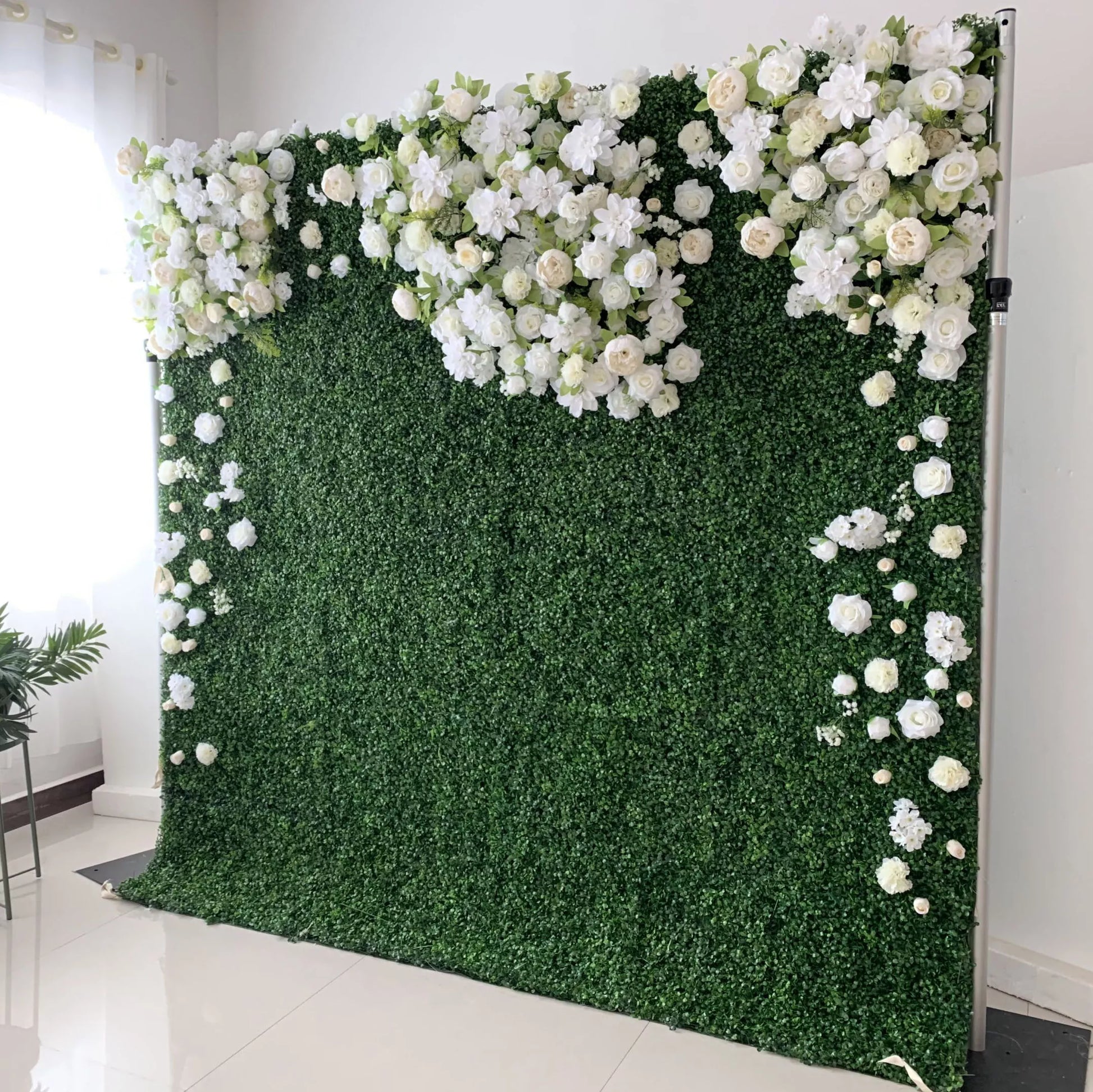 This elegant indoor floral wall features a lush, deep green foliage base, adorned with a cascading arrangement of white and cream blooms—including roses, hydrangeas, and delicate filler flowers. The blooms form a soft, heart-shaped design at the top, with individual flowers trailing down the sides. Set against a minimalist white backdrop, the design evokes a fresh, romantic, and naturally sophisticated atmosphere ideal for weddings, photo backdrops, or elegant celebrations.