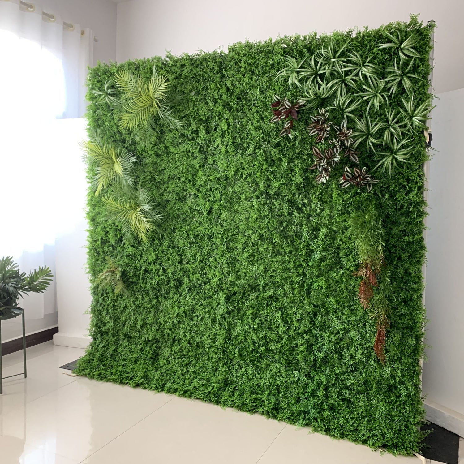This image showcases a lush artificial greenery wall set indoors on a glossy white floor. It features a dense base of vibrant green foliage, artfully accented with varied plants like palm fronds, spiky greens, and variegated leaves. Natural light streams in from a window, enhancing the realistic textures and rich green hues. The diverse elements create a natural, refreshing display, perfect for adding organic elegance to interiors, exuding a serene and sophisticated atmosphere.
