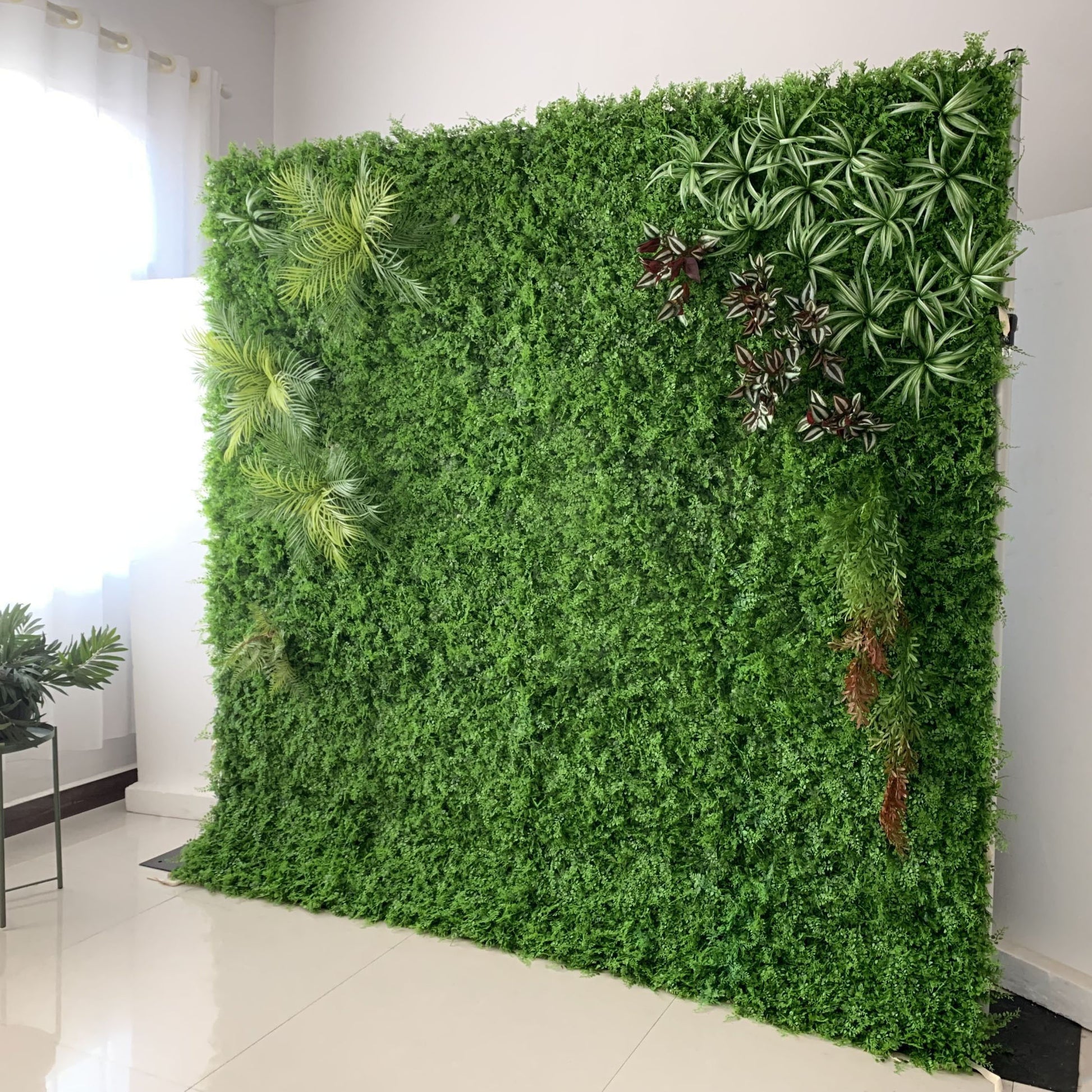 This image showcases a lush artificial greenery wall set indoors on a glossy white floor. It features a dense base of vibrant green foliage, artfully accented with varied plants like palm fronds, spiky greens, and variegated leaves. Natural light streams in from a window, enhancing the realistic textures and rich green hues. The diverse elements create a natural, refreshing display, perfect for adding organic elegance to interiors, exuding a serene and sophisticated atmosphere.