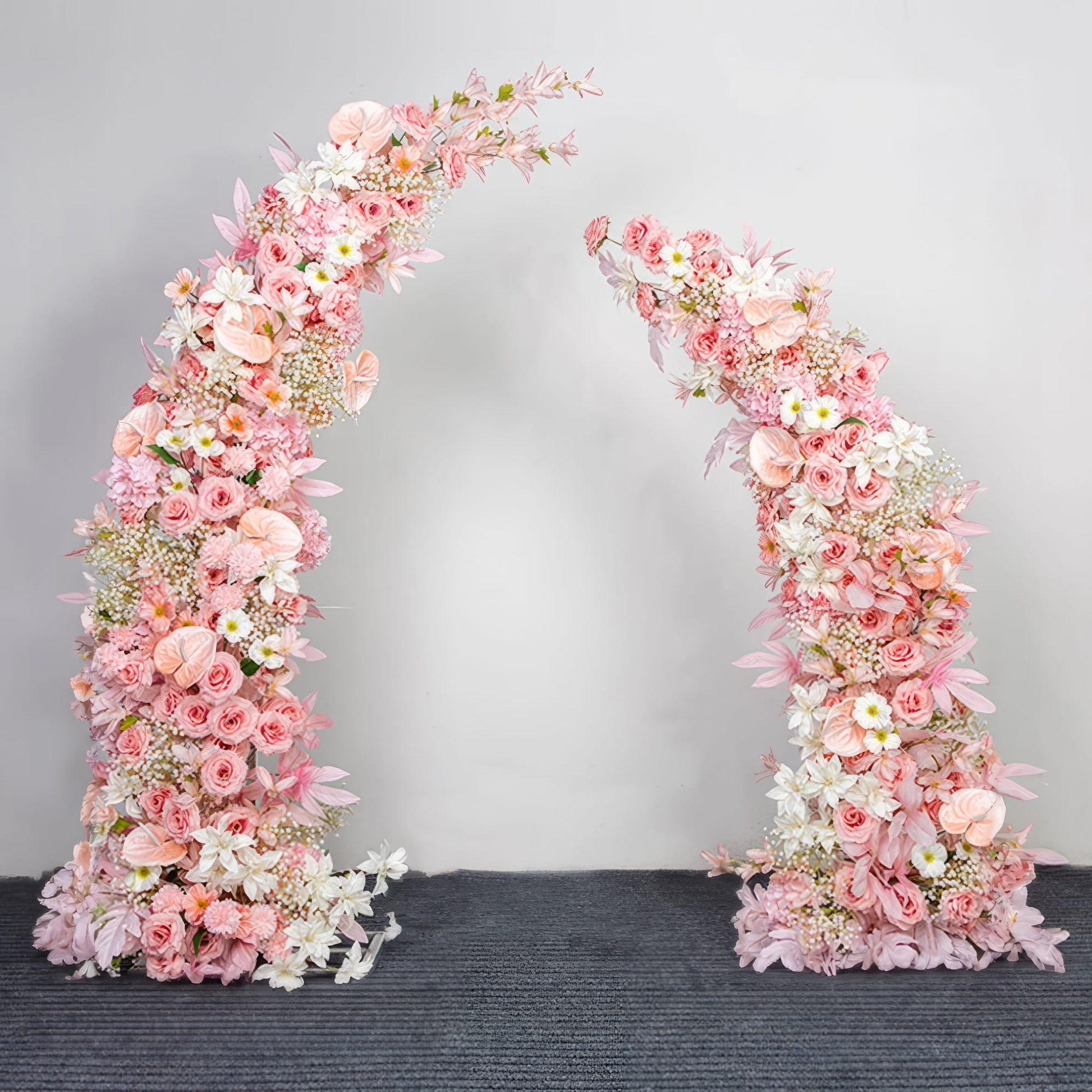 This elegant floral arch from Valar Flowers is a lush, cascading arrangement of soft pink, blush, and white blooms—including roses, peonies, anthuriums, and delicate sprigs—interwoven with vibrant green foliage. Set against a neutral backdrop, it creates a romantic, sophisticated atmosphere ideal for weddings, engagement photoshoots, or elegant celebrations.