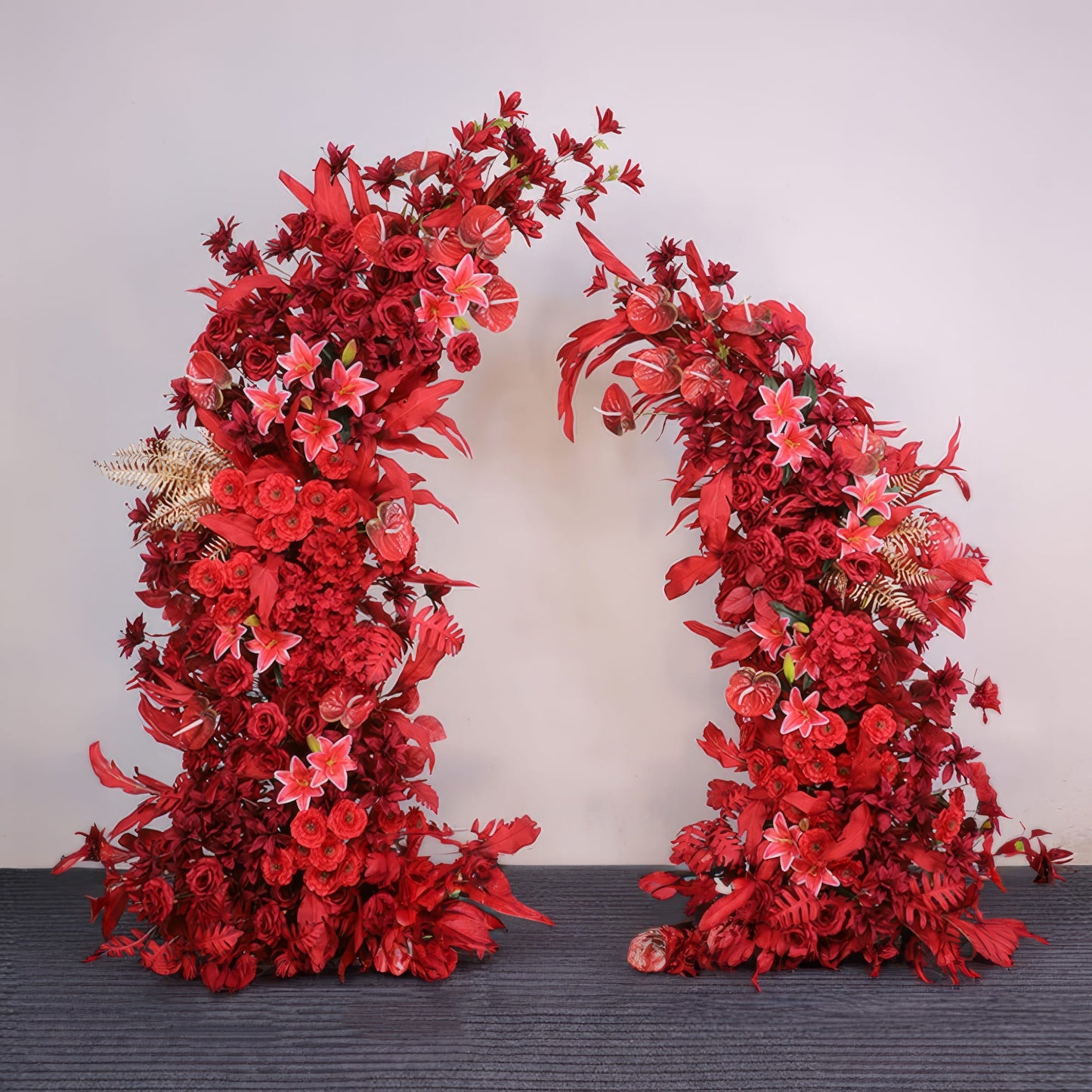 This striking floral arch from Valar Flowers is a bold, cascading arrangement of vibrant red blooms—including roses, lilies, and anthuriums—interwoven with textured foliage and subtle gold accents. Set against a neutral backdrop, it creates a dramatic, passionate atmosphere ideal for weddings, engagement photoshoots, or unforgettable celebrations.