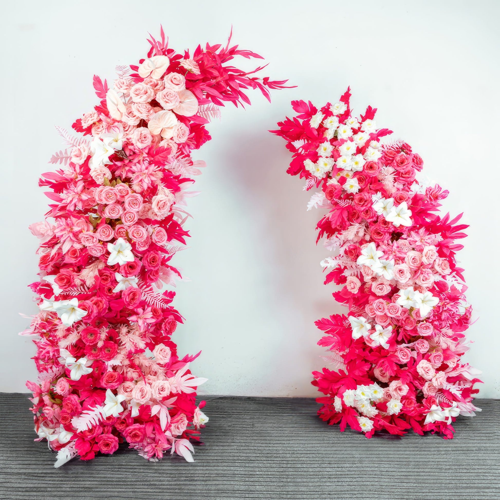 This vibrant floral arch from Valar Flowers is a lush, cascading arrangement of soft pink, deep fuchsia, and white blooms—including roses, lilies, and delicate sprigs—interwoven with bold red foliage. Set against a neutral backdrop, it creates a romantic, energetic atmosphere ideal for weddings, engagement photoshoots, or joyful celebrations.