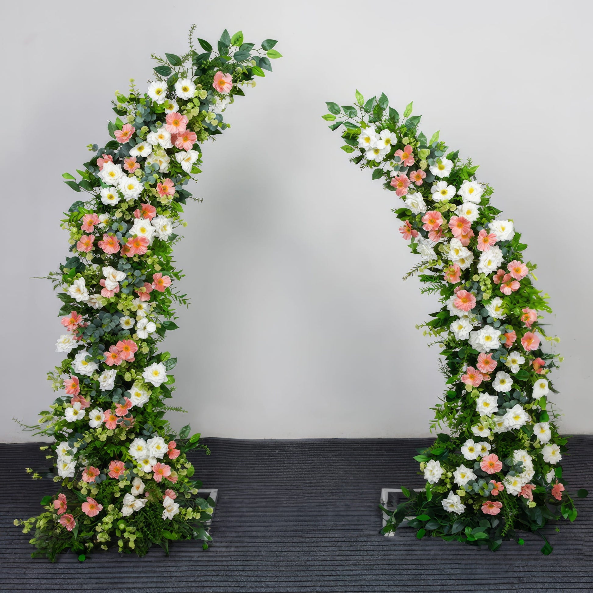 A romantic, asymmetrical floral arch from Valar Flowers, crafted with a fresh palette of blush pink, creamy white, and vibrant green blooms—including roses, daisies, and lush foliage. Set against a neutral studio backdrop, it creates a joyful, elegant, and eye-catching aesthetic ideal for wedding ceremonies, photo sessions, and special event decor.