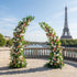 A romantic, asymmetrical floral arch from Valar Flowers, crafted with a fresh palette of blush pink, creamy white, and vibrant green blooms—including roses, daisies, and lush foliage. Set on a sunlit Parisian riverside terrace with the iconic Eiffel Tower in the background, it creates a joyful, elegant, and picturesque atmosphere perfect for luxury weddings and editorial photoshoots.