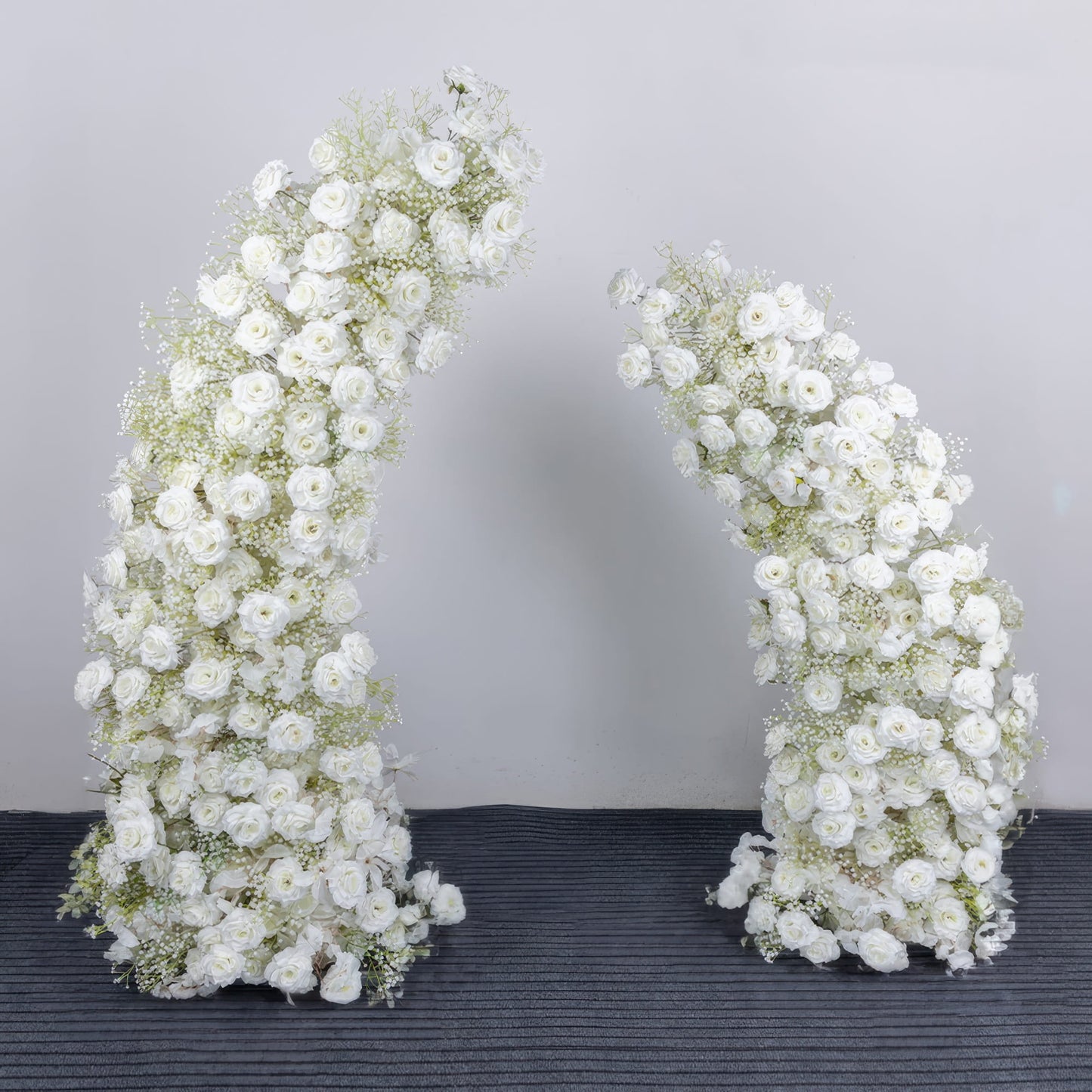 A timeless, asymmetrical floral arch from **Valar Flowers**, crafted with a pure white palette of lush roses, peonies, and delicate baby's breath. Set against a neutral studio backdrop, it creates a serene, elegant, and ethereal aesthetic ideal for wedding ceremonies, photo shoots, or special event decor.
