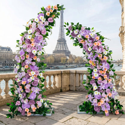 A vibrant, asymmetrical floral arch from Valar Flowers, bursting with a lively blend of lavender, soft pink, peach, and white blooms—including roses, peonies, and daisies—intertwined with lush greenery. Set on a sunlit Parisian riverside terrace with the iconic Eiffel Tower in the background, it creates a joyful, romantic, and picturesque aesthetic ideal for luxury weddings and editorial photoshoots.