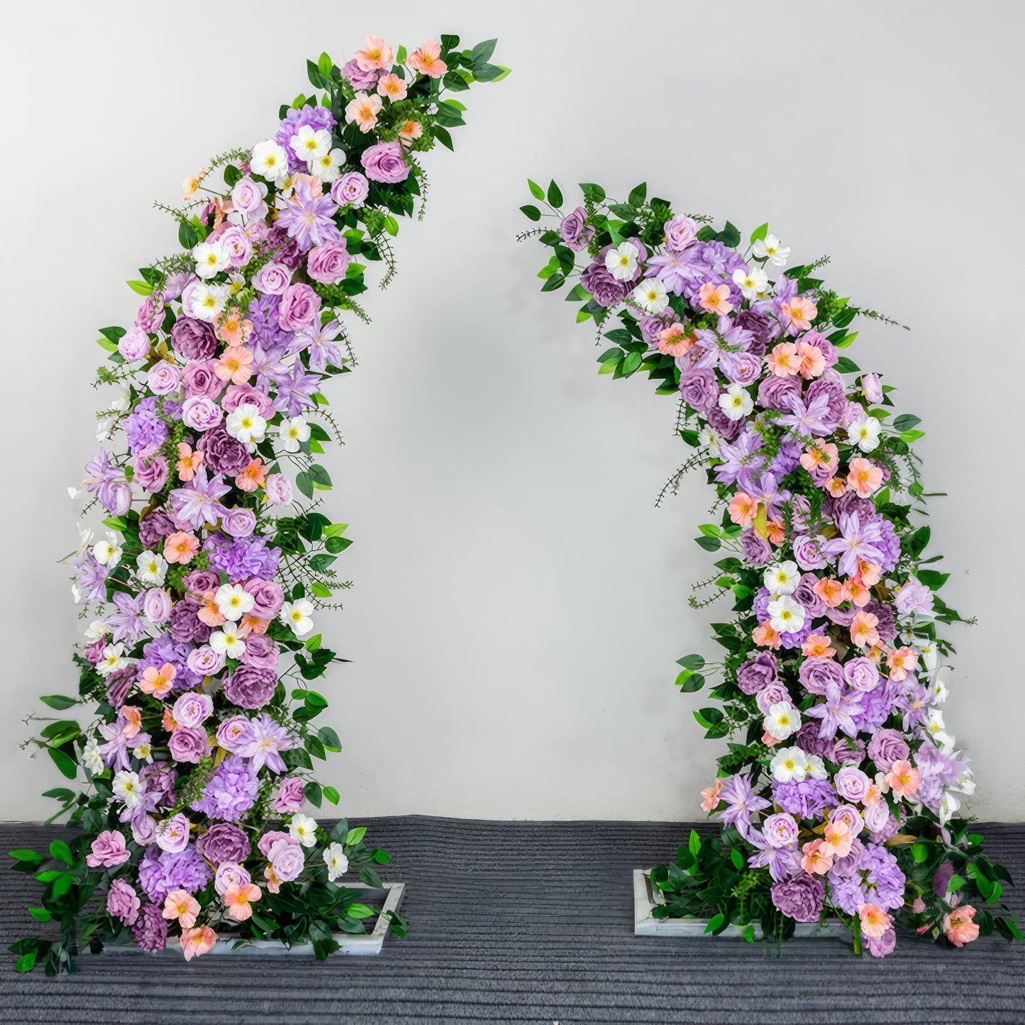 This stunning floral arch from Valar Flowers features a vibrant blend of lavender purple, soft peach, and creamy white blooms—including roses and peonies—intertwined with lush green foliage. The asymmetrical design, set against a plain backdrop, creates a romantic and eye-catching aesthetic perfect for wedding ceremonies, photo sessions, and special event decor.