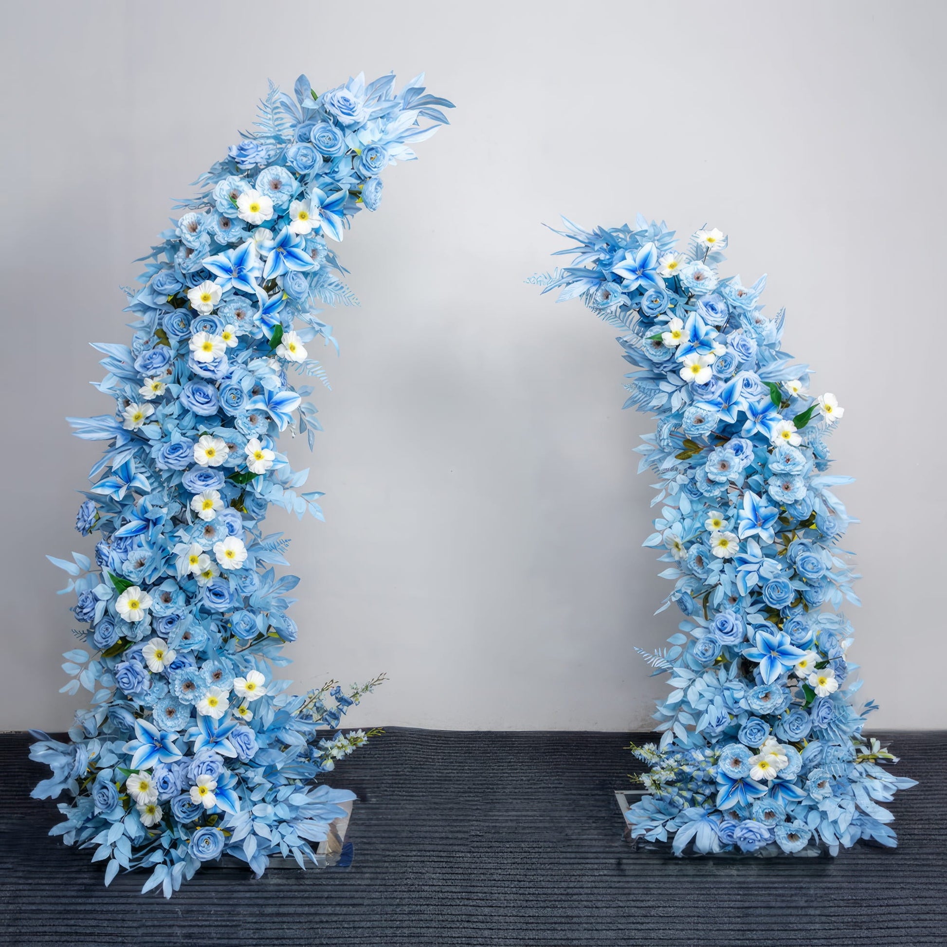 This is a stunning, asymmetrical floral arch from **Valar Flowers**, crafted with a serene palette of sky-blue roses, lilies, and hydrangeas, accented by delicate white blooms with yellow centers. Set against a neutral studio backdrop, it creates a cool, elegant, and dreamy aesthetic ideal for weddings, photo shoots, or special event decor.