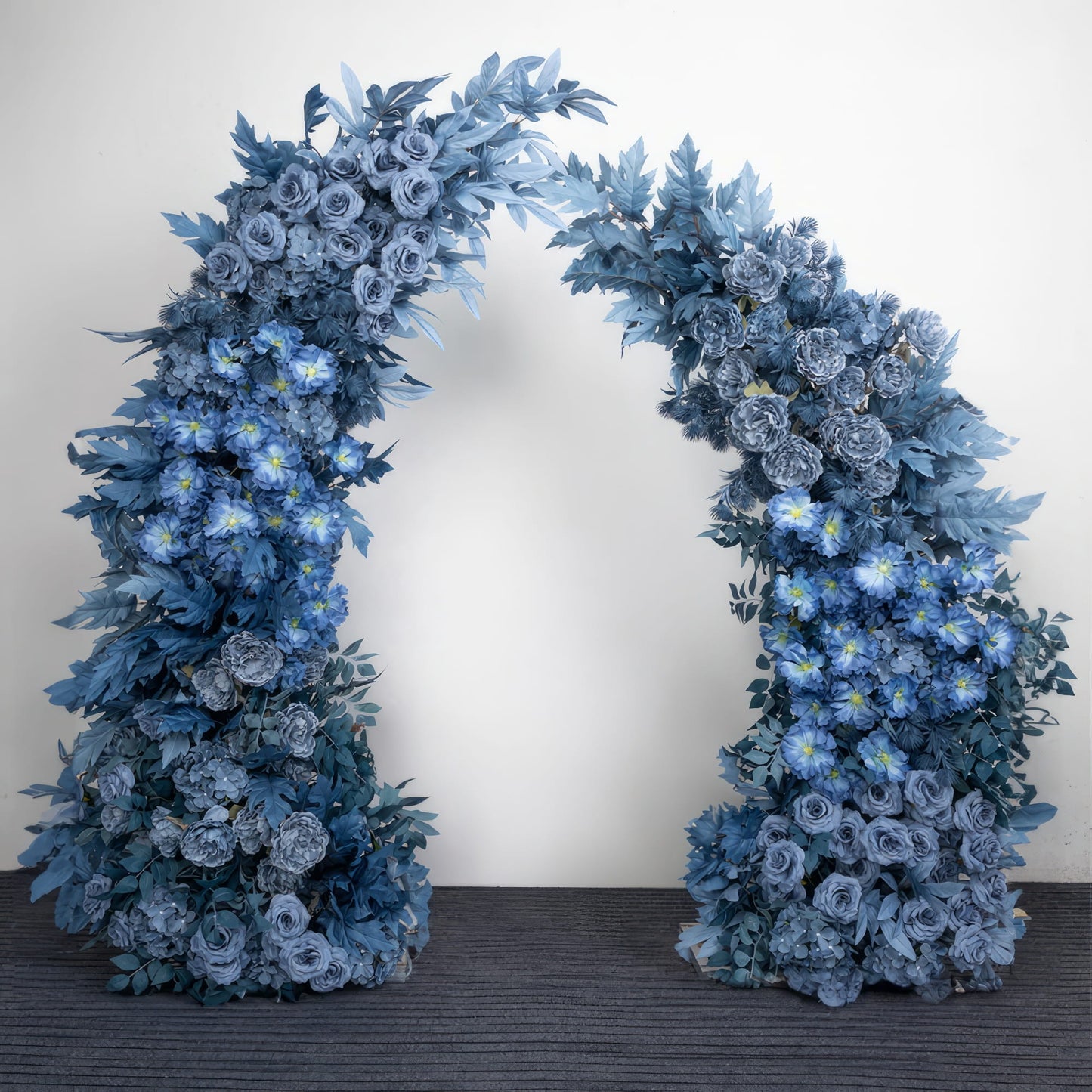 Stunning blue floral archway featuring gradient hues of denim, sky blue, and navy, crafted from lush roses, peonies, and daisies interwoven with textured greenery. Set against a minimalist studio backdrop, it creates a serene, sophisticated atmosphere ideal for wedding ceremonies, photoshoots, and special event decor.