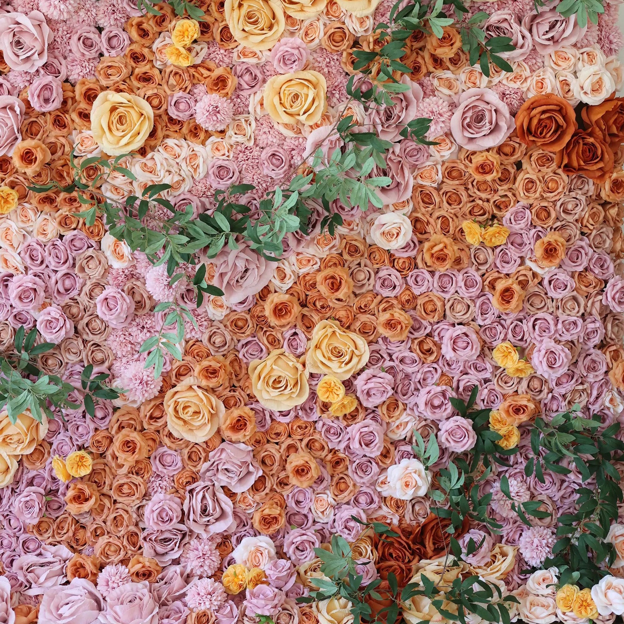 This image showcases a stunning floral wall by Valar Flowers. It features a dense, artful arrangement of artificial roses in a rich palette of soft pinks, warm oranges, and creamy yellows, interspersed with lush green foliage. The flowers create a lush, textured, and visually captivating display, exuding elegance and charm, perfect for adding a touch of sophistication to events or interior spaces.