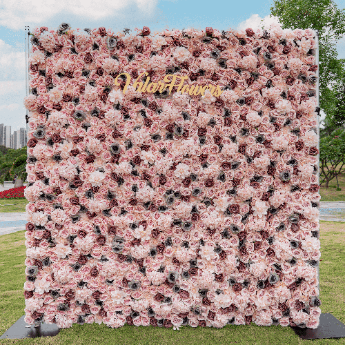 This flower wall is densely packed with pink-hued roses, each accentuated with dark, intricate details in their centers. The uniform design offers a sumptuous visual texture. A golden "Valar Flowers" sign crowns the top, contrasting the blooms. Positioned outdoors, it stands as a vivid focal point against a city skyline backdrop.