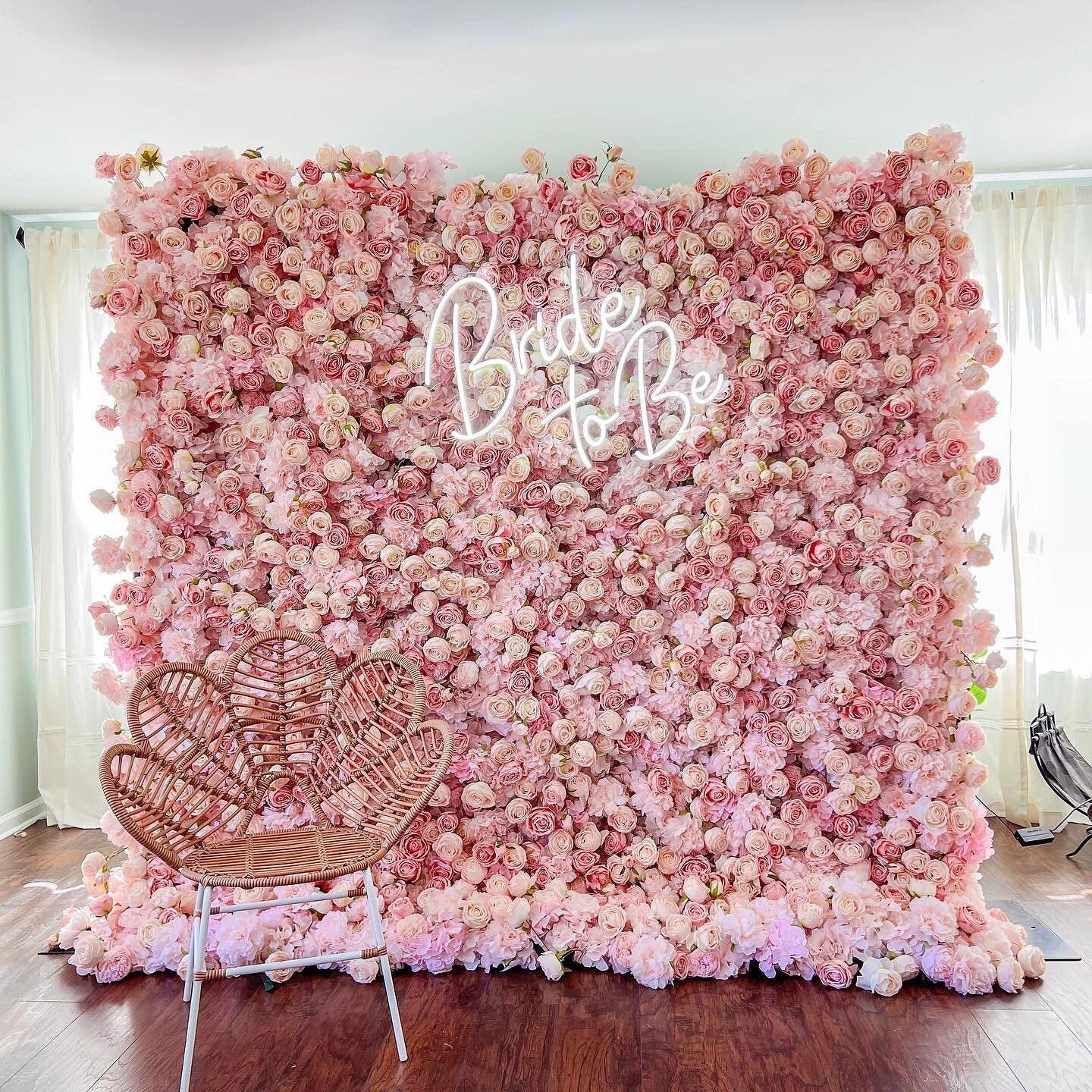 A beautiful flower wall densely covered with pink roses. In the center, neon lights spell out &quot;Bride To Be&quot;. A wicker chair sits in front, creating a romantic bridal setting.