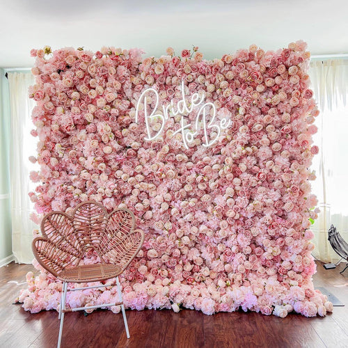 This photo showcases a stunning floral backdrop densely covered in soft pink roses. The word “Bride to Be” is prominently displayed in elegant neon lights, adding a celebratory touch. In the foreground, a heart - shaped wicker chair with a woven design sits on a dark wooden floor, complementing the romantic setting. Softly lit by natural light filtering through sheer curtains, the scene exudes charm and is perfect for bridal events. 